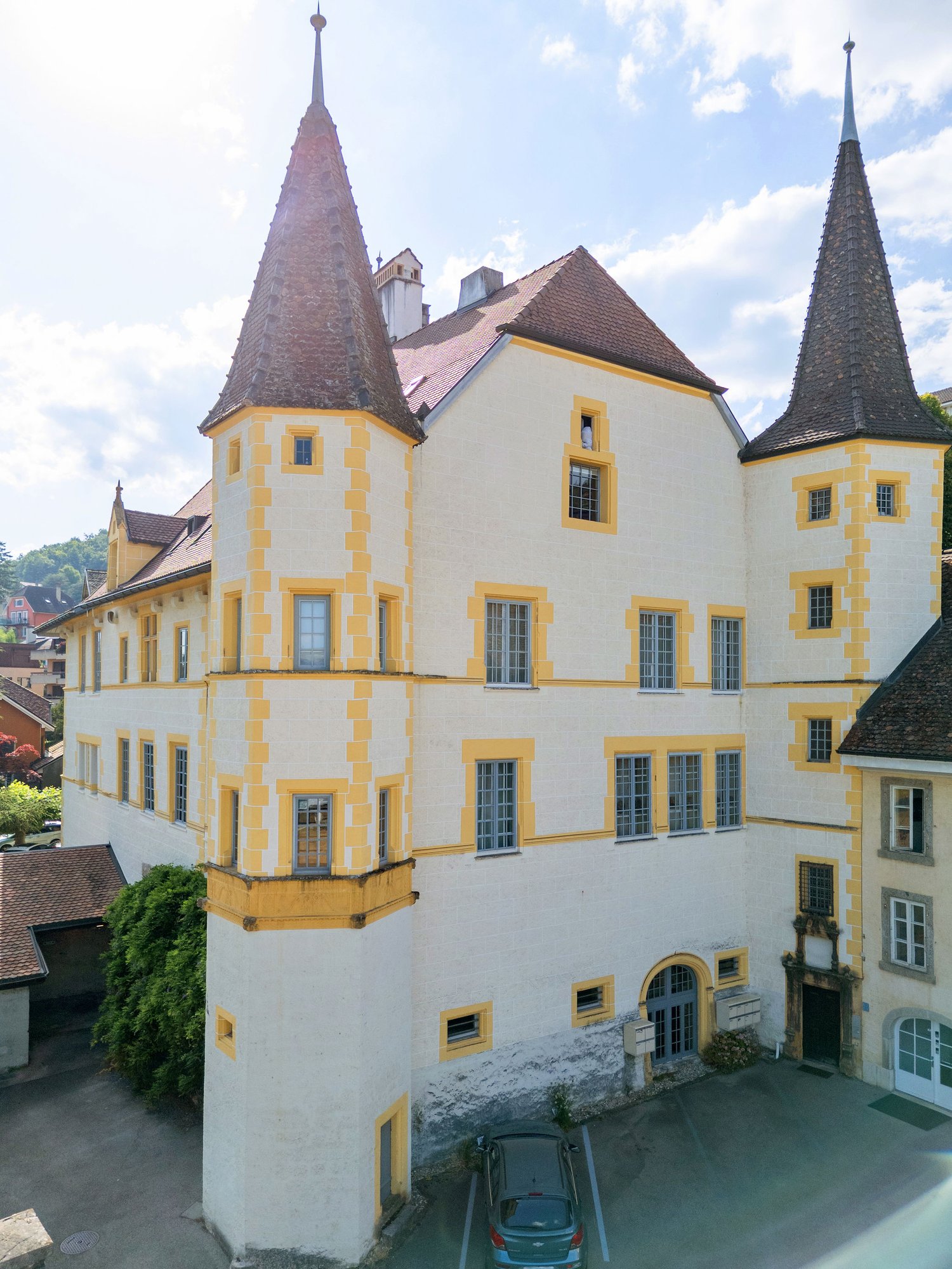 Large white building with yellow trim, two towers, multiple windows, brown roof, car parked in front