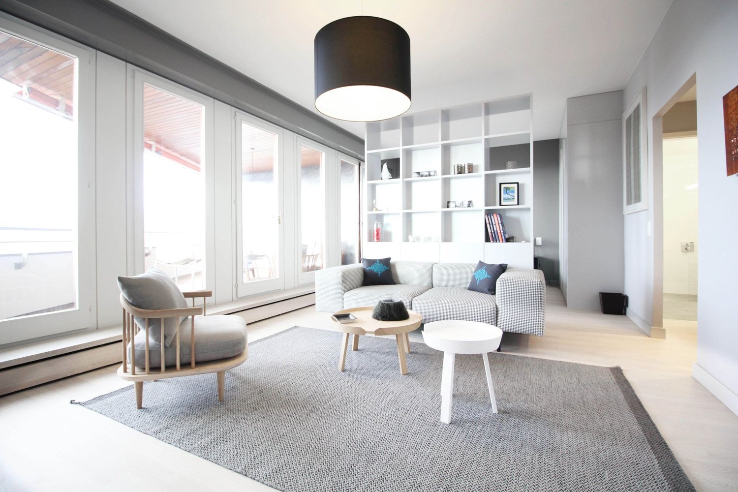 modern living room with white walls, large windows, wooden floor, gray rug, white couch, wooden chair, white coffee table, black pendant light