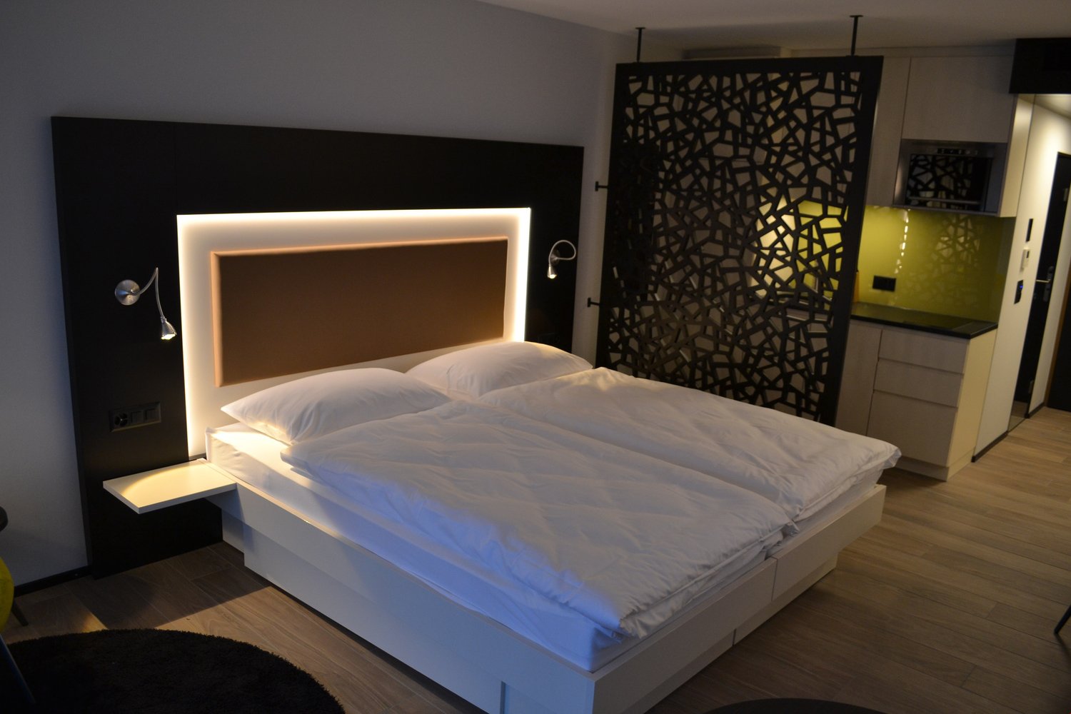 Modern bedroom with a large bed, white bedding, and a decorative black and white patterned wall panel. The room also features recessed lighting in the headboard and a fireplace or heating unit on the wall.