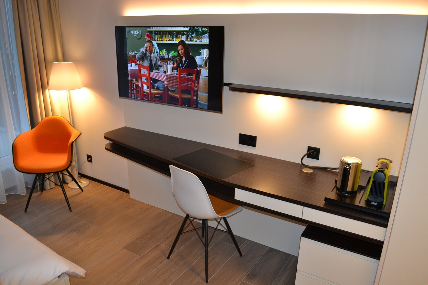 Modern living room with a large TV mounted on the wall, a dark wooden TV stand with drawers, an orange accent chair, and a white chair at a desk area. The room has hardwood flooring and appears to have a balcony or terrace visible through the window.