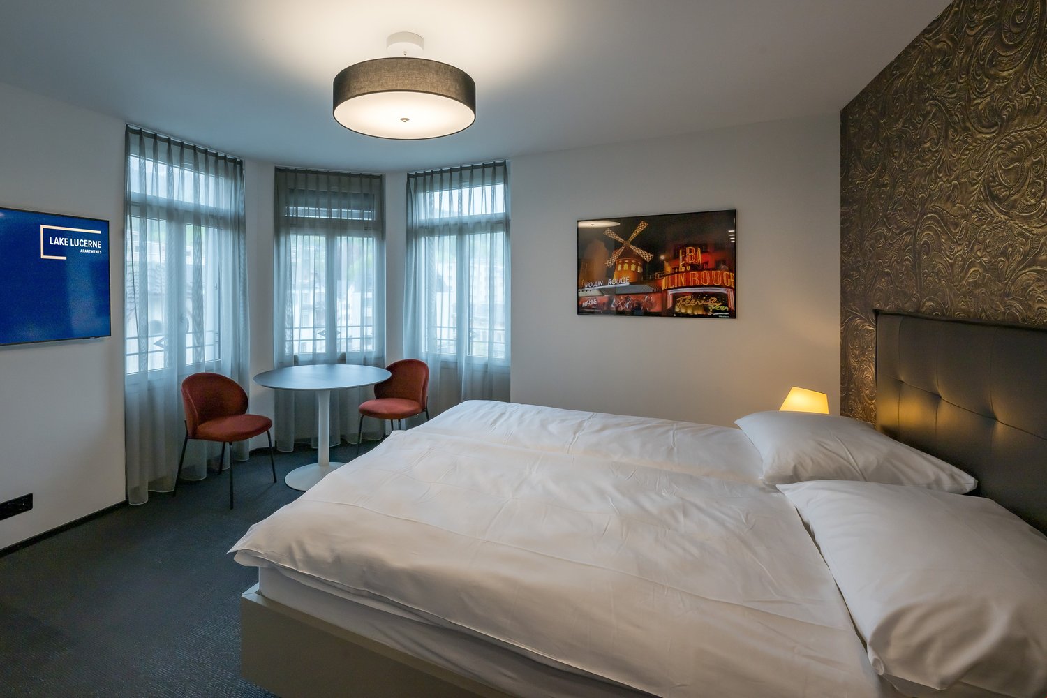 Single bed, round table, two chairs, curtained windows, lamp, mounted picture, flat screen TV