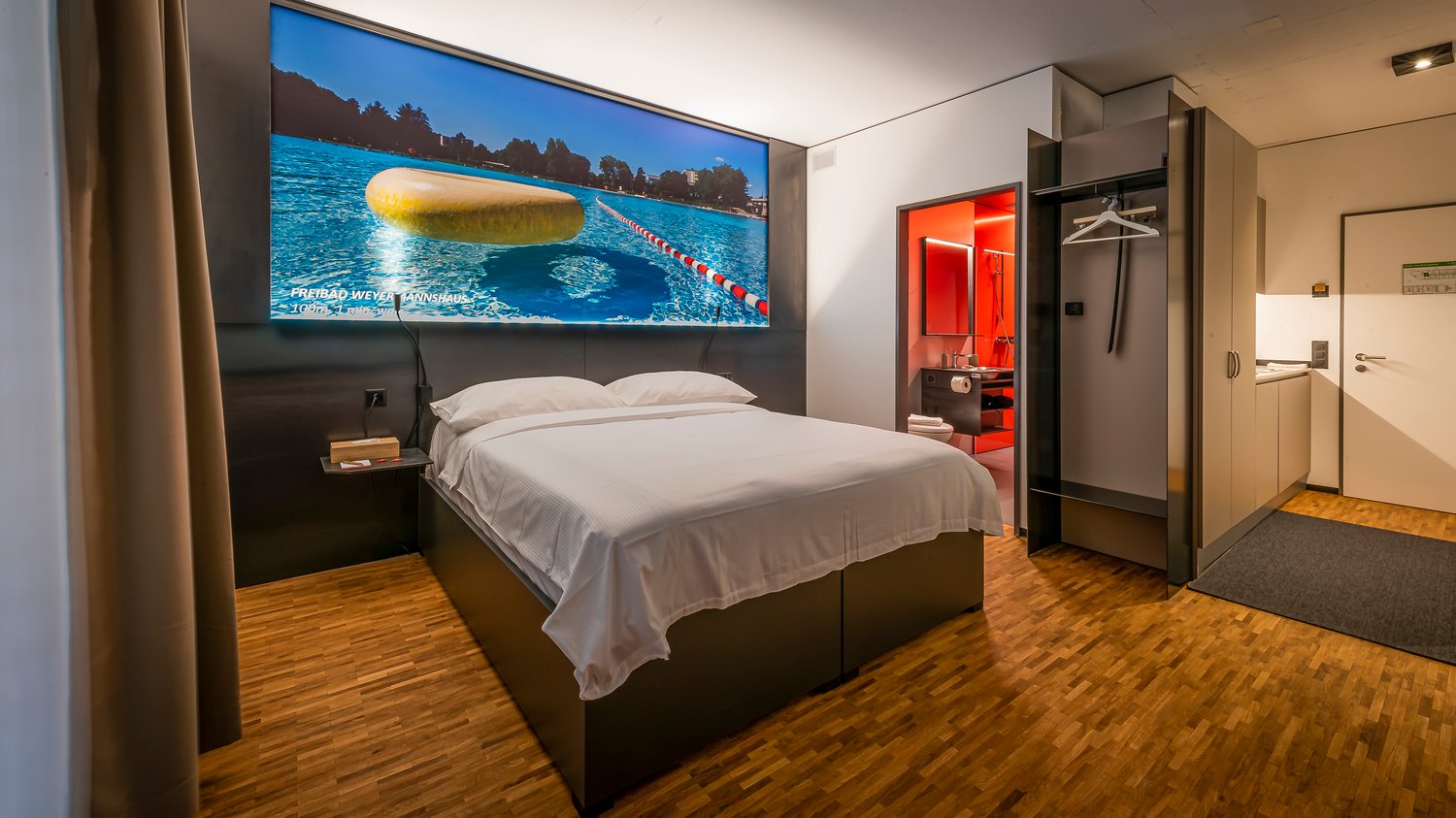 Large bedroom with a king-size bed, hardwood flooring, and a large wall-mounted TV displaying a scenic image of a lake and mountains. The room also has a closet and a bathroom with a red accent wall.