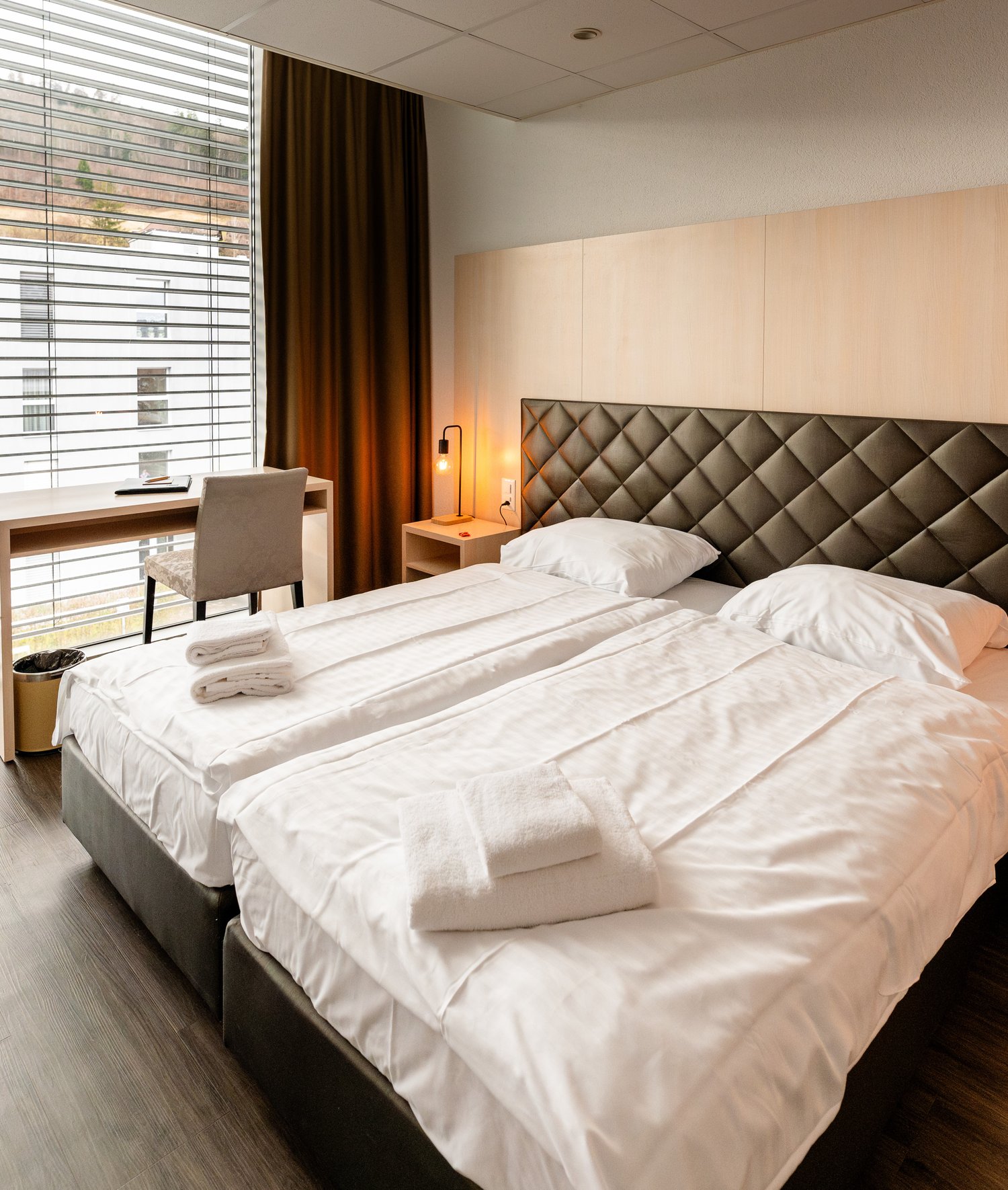 Spacious bedroom with large windows, quilted headboard, bedside tables, and a desk area