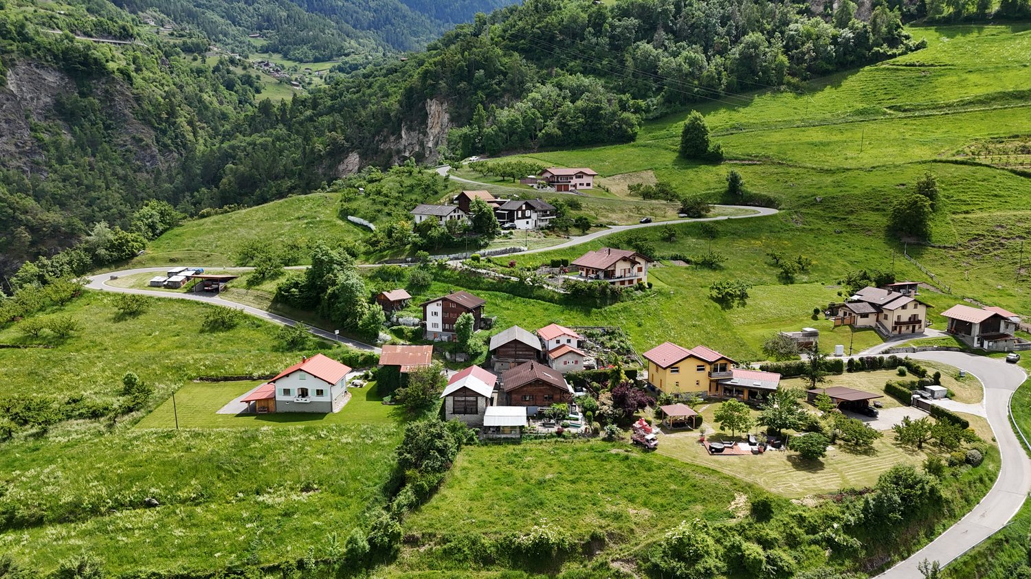 Aerial view of a picturesque mountain village nestled in a lush, green valley. The village features a cluster of traditional alpine-style houses with red roofs, surrounded by rolling hills, forests, and rocky cliffs. A winding road runs through the villag