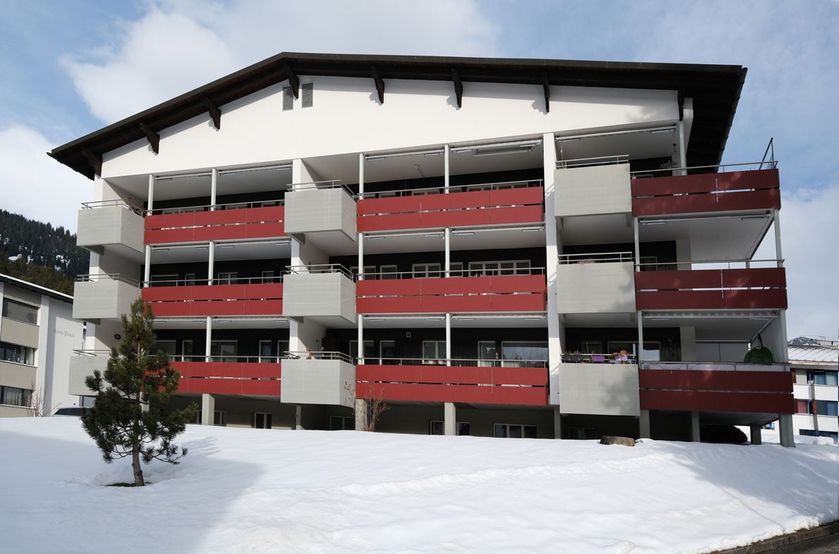 Multi-story building, white exterior, red balconies, covered balconies, located in snowy area, front garden