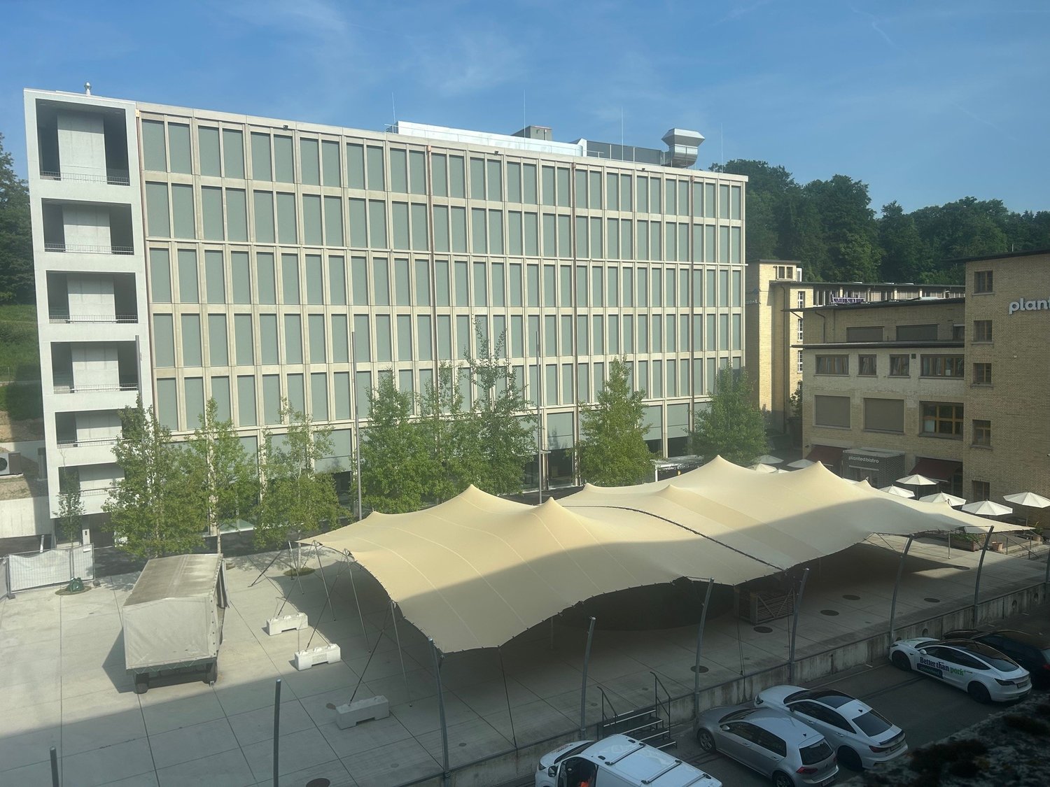 Multi-story office building with glass facade, outdoor tents set up in the plaza area, and parking spaces visible in the foreground.