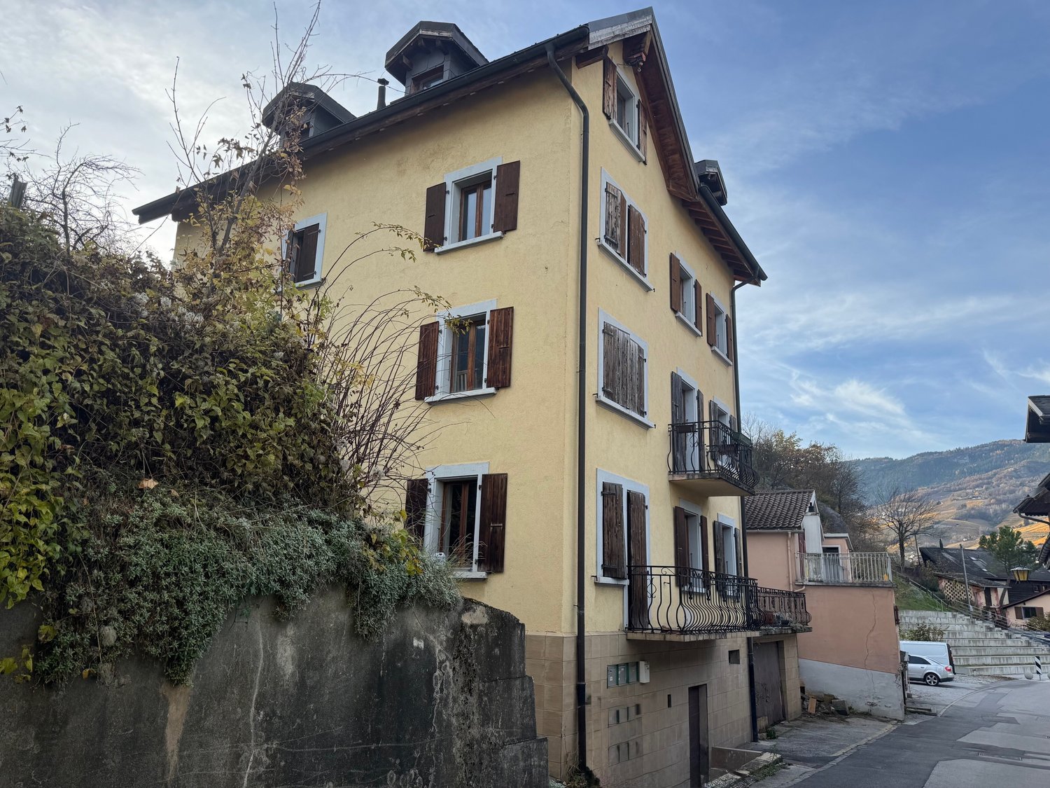 A three-story yellow building with shuttered windows and two balconies, situated on a hill with a concrete wall and greenery.