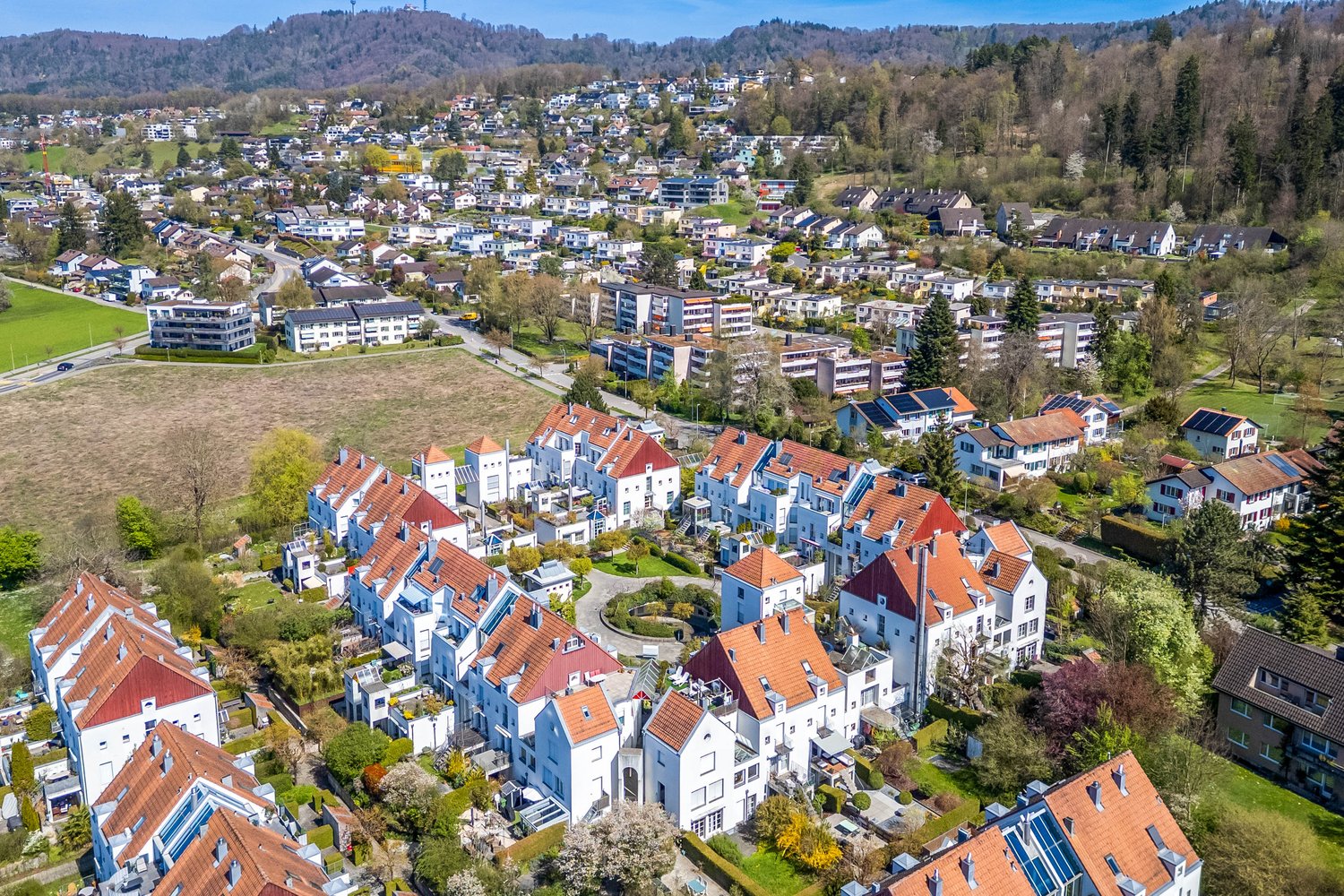 Aerial view of a residential area with many houses, some with red roofs, surrounded by trees and greenery.