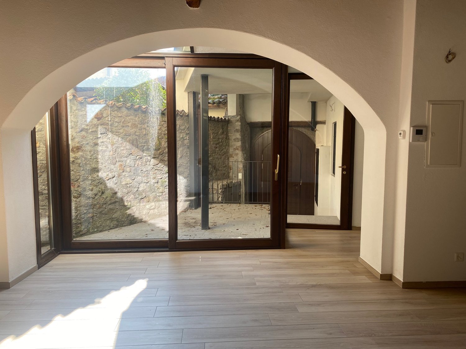The image shows a spacious interior with an arched entryway leading to a balcony or terrace area. The room has hardwood flooring and a stone wall visible through the glass doors. The interior appears to be well-lit with natural light coming in through the