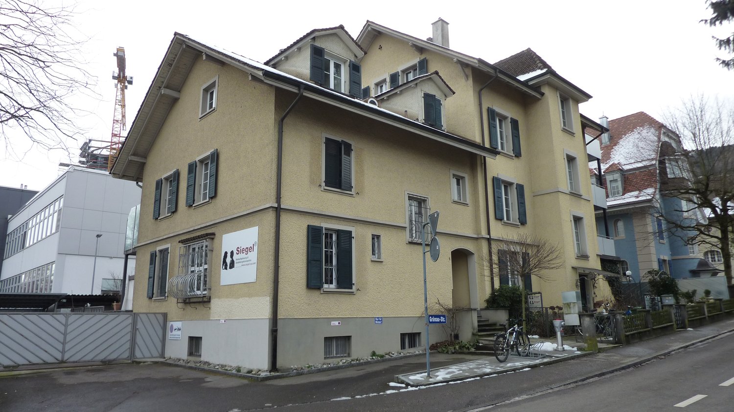 Yellow facade building, several floors, balconies, windows with shutters, sign with 'Siegel' on the wall, bicycles parked on the sidewalk