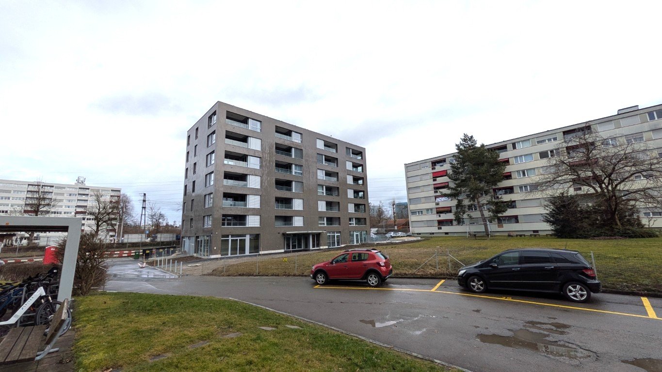 modern building, glass windows, grey walls, two cars parked, bicycles near a railing, grass and trees