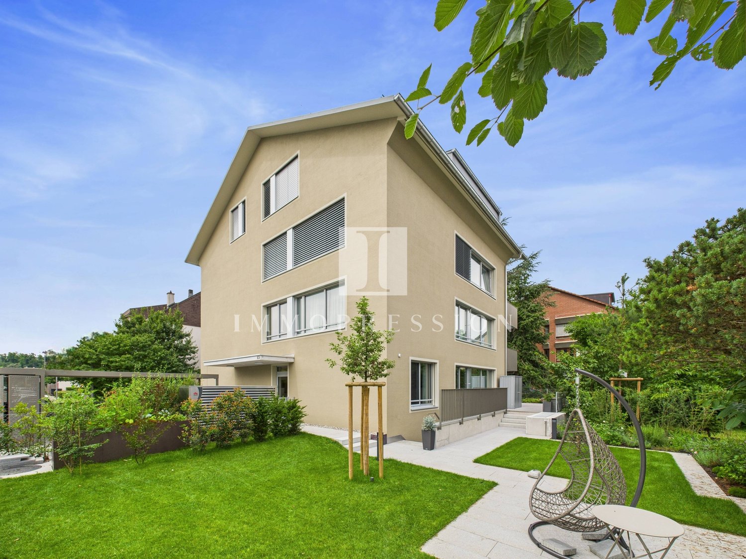 Modern 2-story house, beige exterior, large windows, well-maintained garden, patio with hanging chair, tree in the center