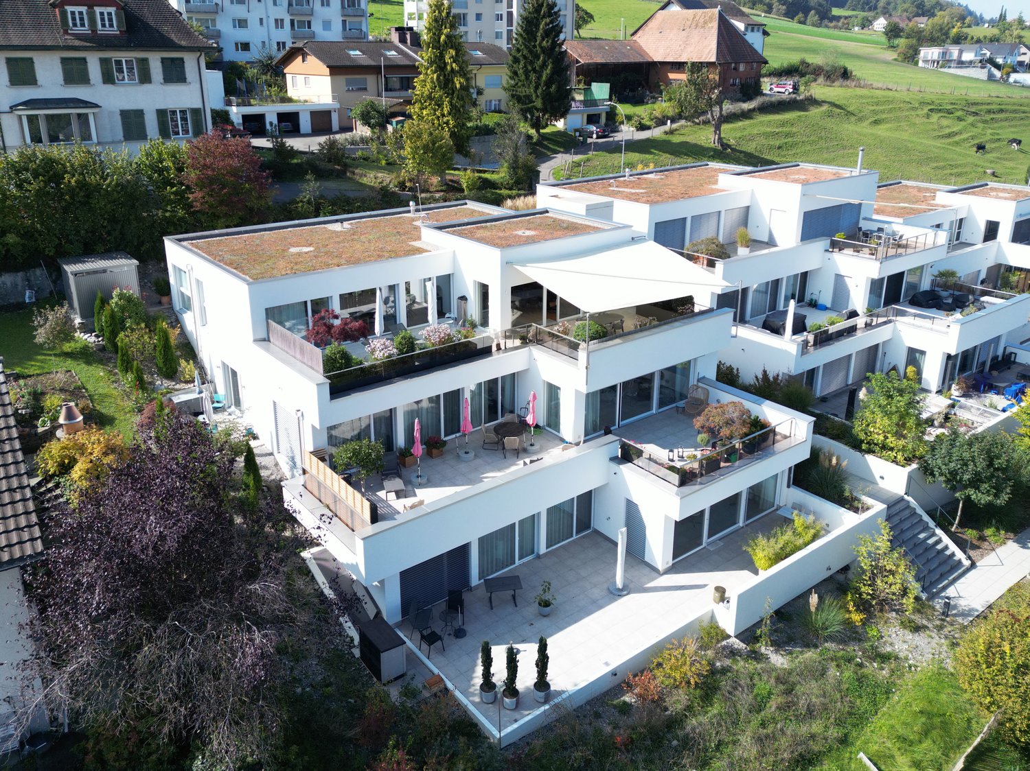 Modern white apartment building, 3 stories, multiple balconies, terrace area with umbrellas, garden areas, view of the surrounding countryside.