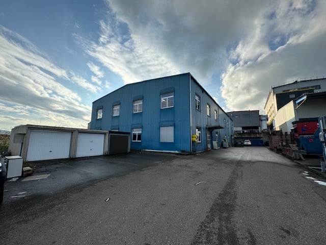 Two-story industrial building with a blue exterior, multiple windows, and garages or parking spaces in the front area