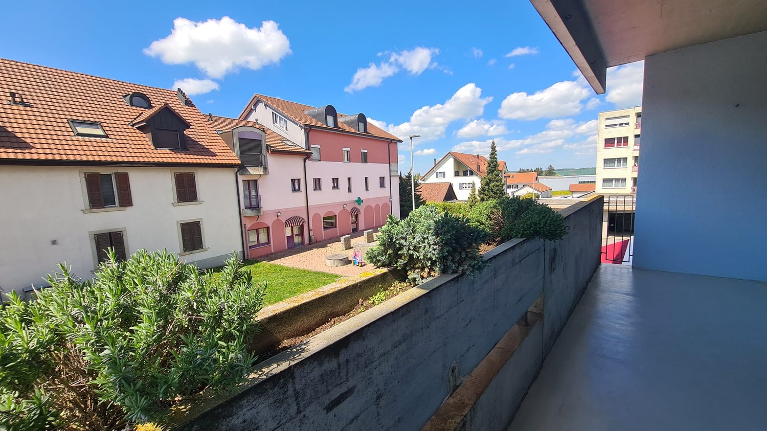 Building with multiple floors, red roof, balcony, concrete wall, plants, street light, houses in the background