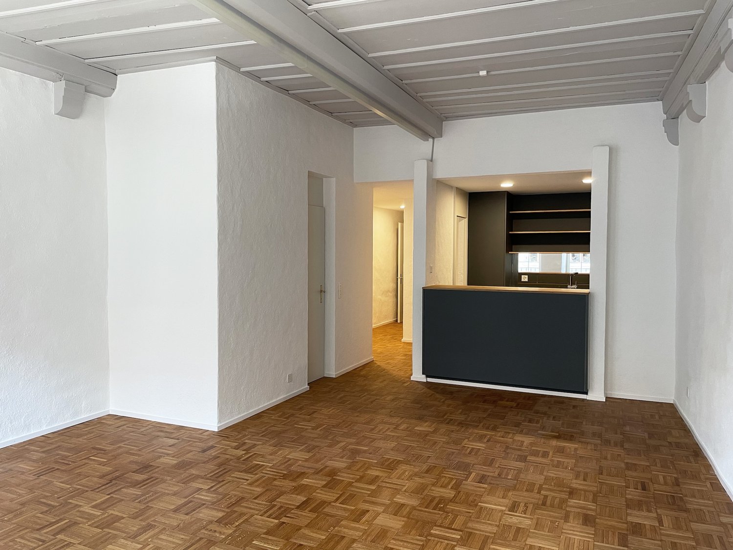Empty room, wooden floor, white walls, ceiling beams, open kitchen with shelves and sink