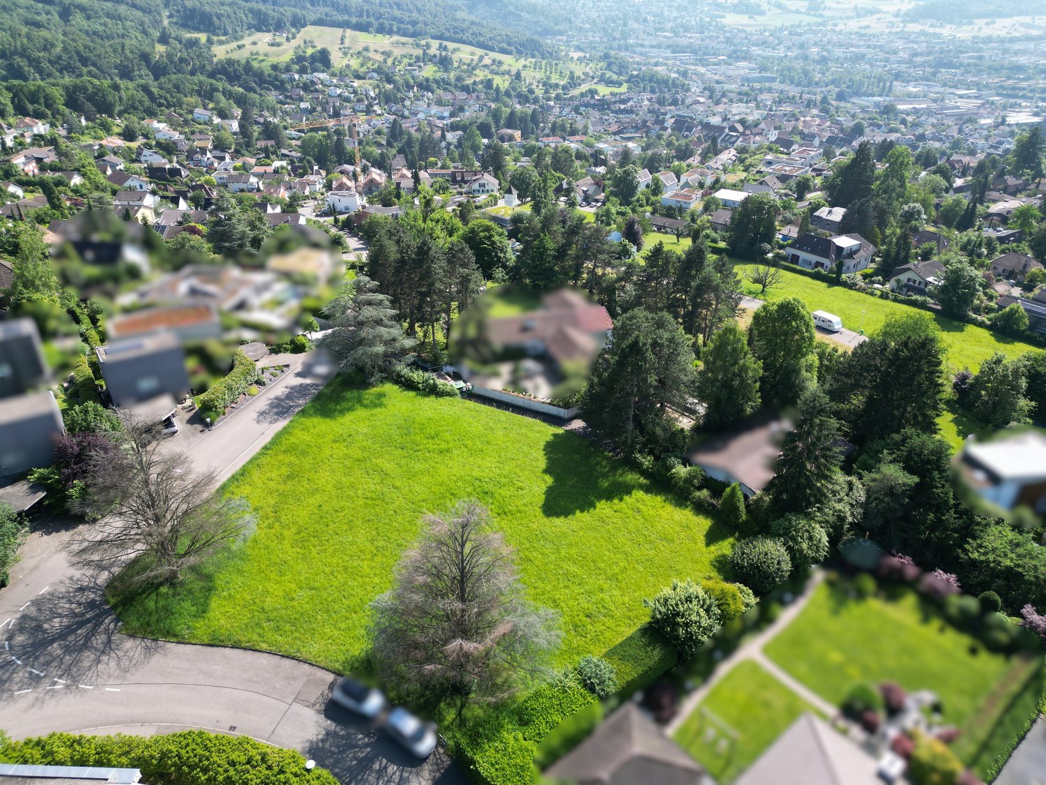 An aerial view of a suburban neighborhood with houses, green fields, and trees. The houses are mostly detached single-family homes with well-kept yards. The area is surrounded by trees, and there are hills in the background. A driveway and car are visible