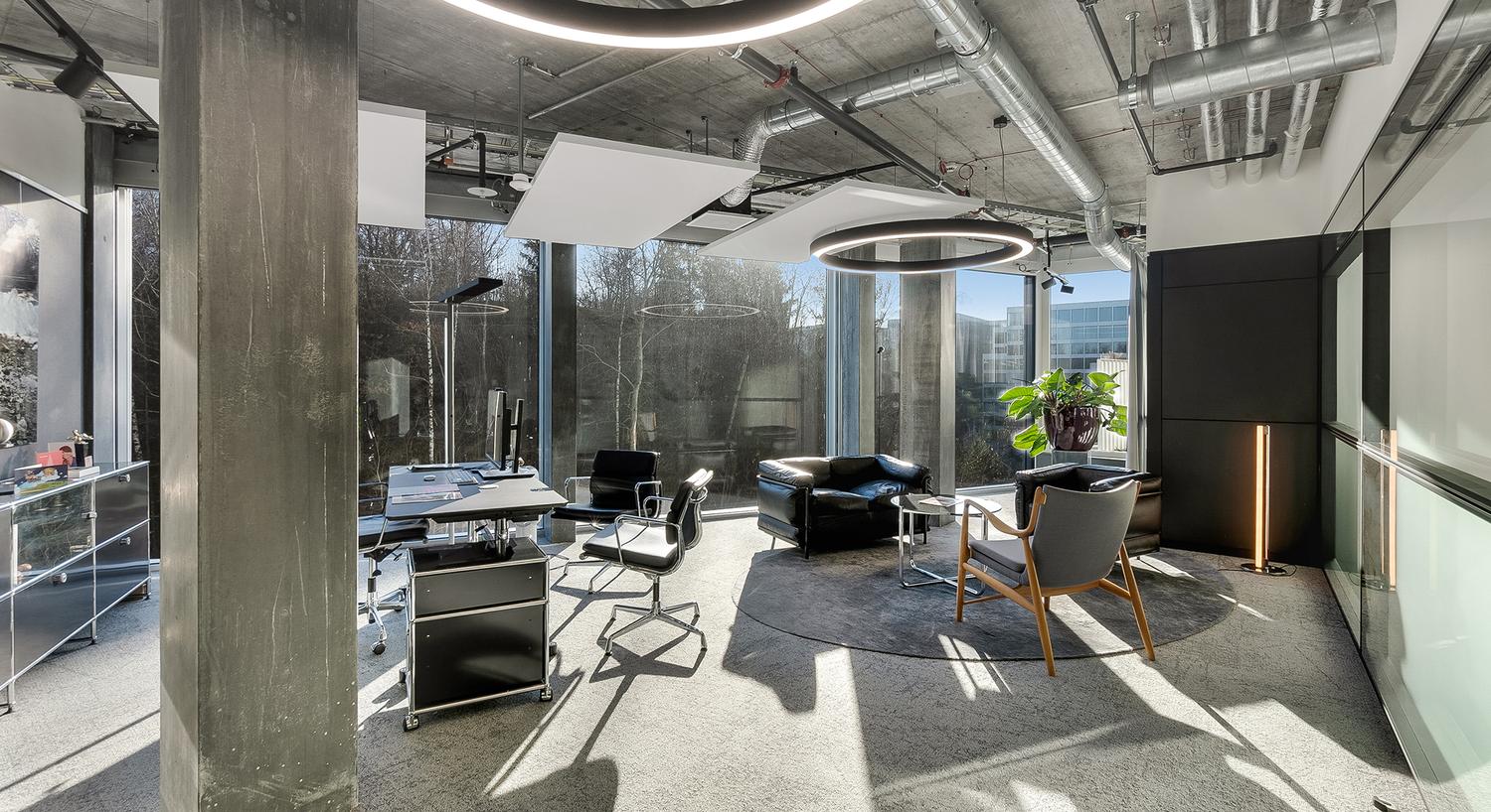 Modern, open-concept office space with concrete walls and ceiling, large windows providing a view of the outdoors, black leather furniture including sofas and chairs, and a central work desk with computer equipment.