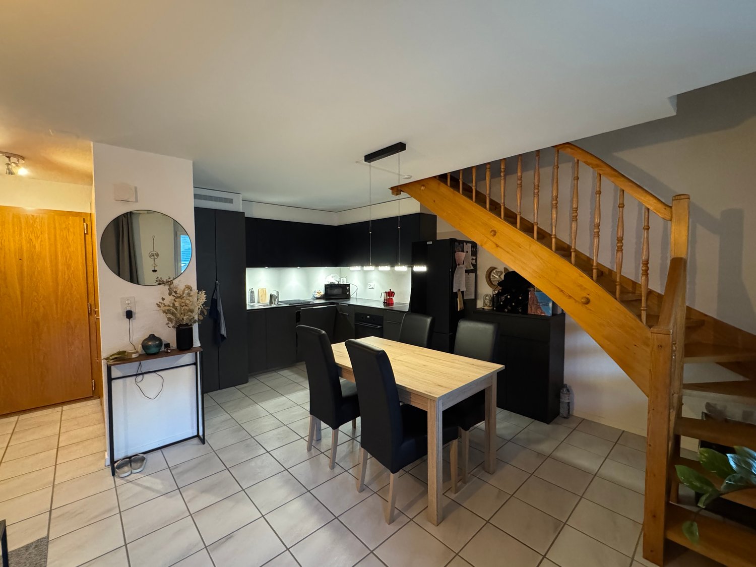 Dining area with a wooden table and black chairs, wooden staircase, kitchen with black cabinets, round mirror, and a wooden door.
