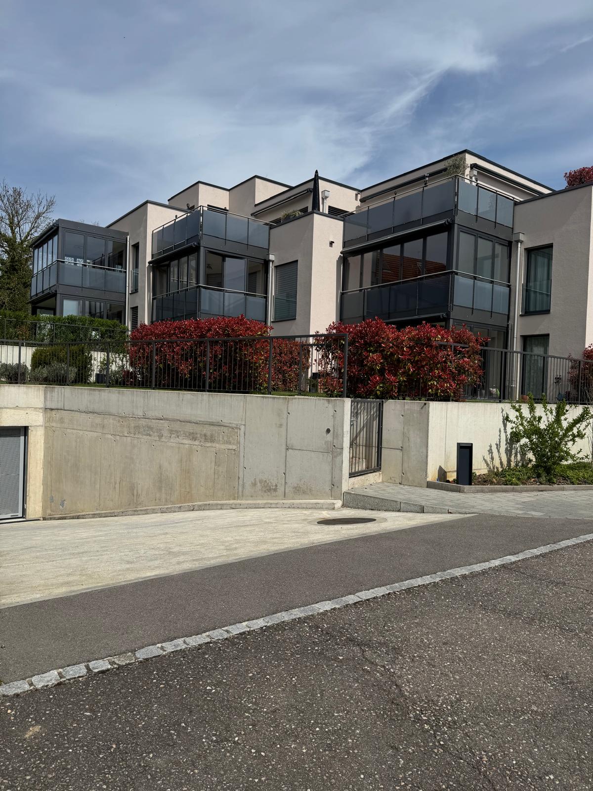 Modern apartment building with glass balconies, concrete wall, black railings, red bushes, and a concrete driveway.