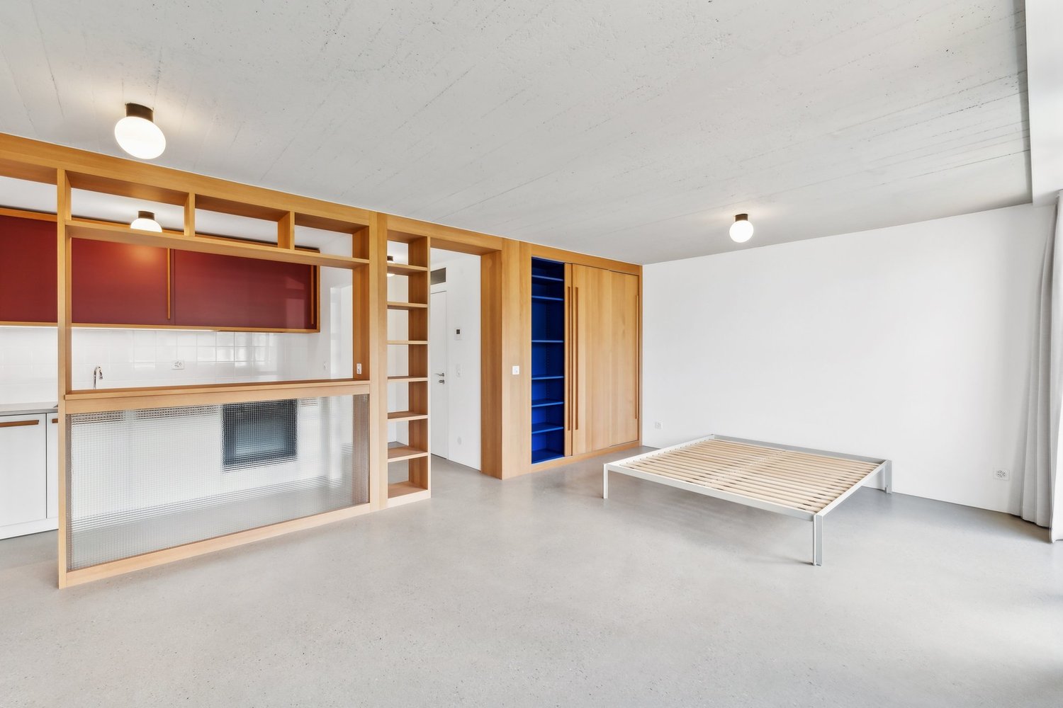 Open-concept studio apartment with wooden shelving units, a kitchenette with a dishwasher, and a raised platform bed area