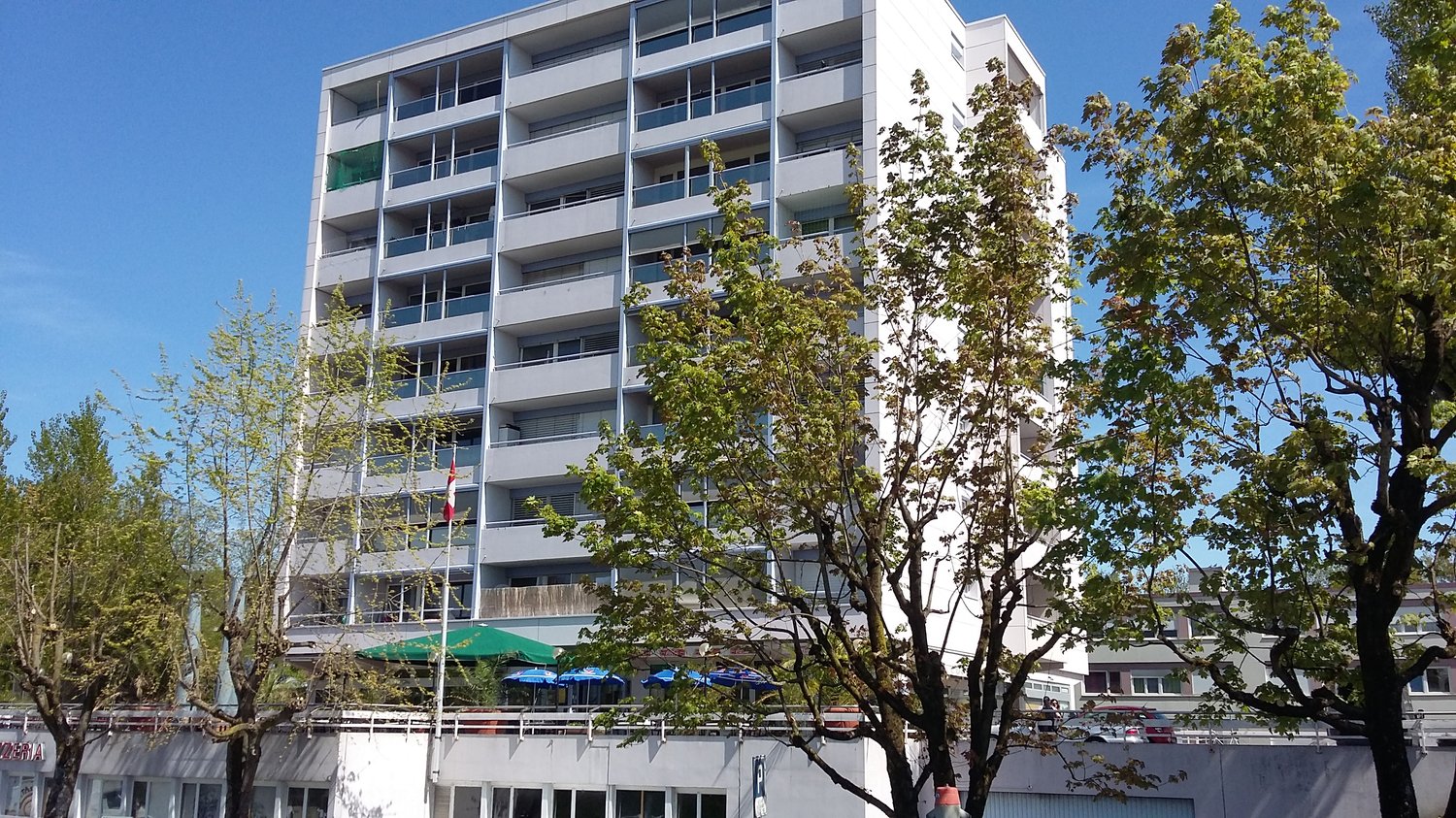 Modern multi-story apartment building with large glass windows, green awnings, a flag pole, and a tree-lined street.