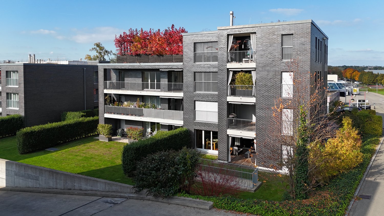 A modern, gray-bricked building with multiple floors, balconies on several levels, and greenery such as hedges and small trees.