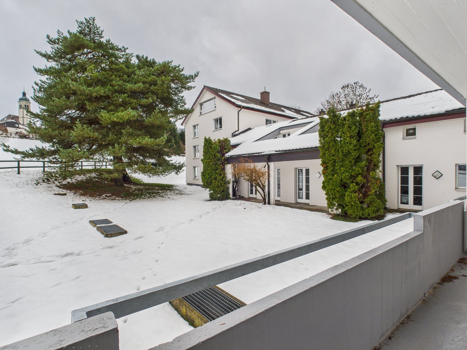 Two-story building, white exterior, covered in snow, large front yard, snow on ground, trees, small solar panels, fenced yard, another building in the background.