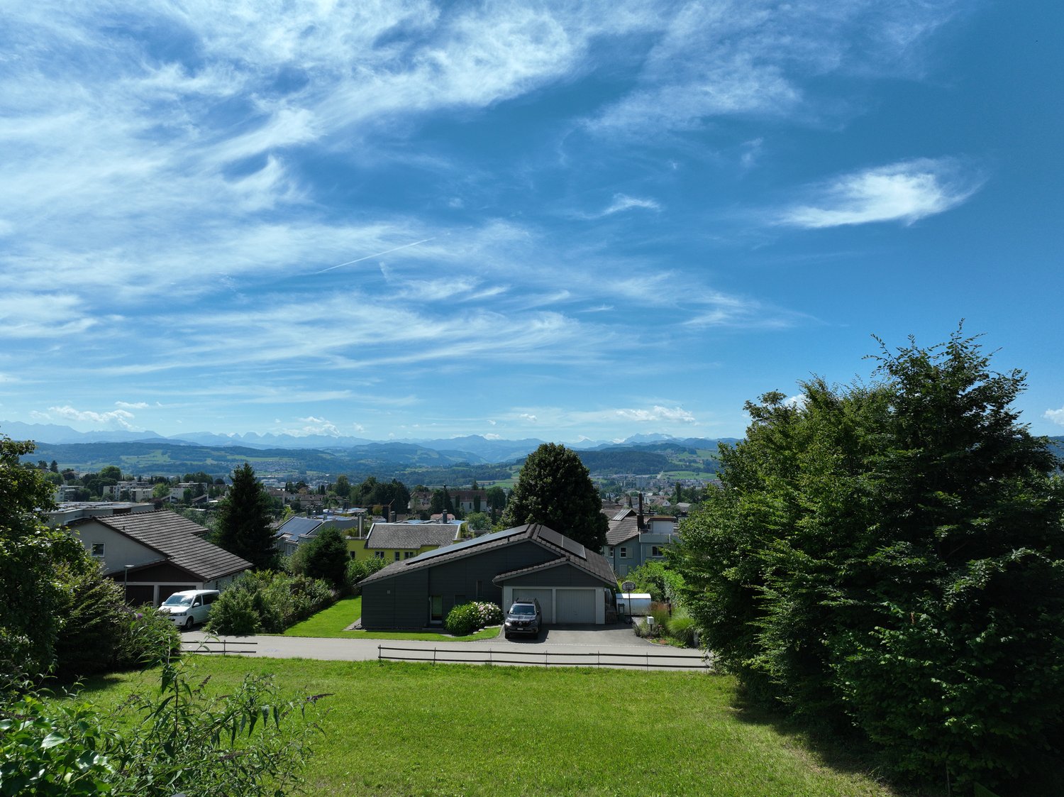 The image shows a scenic view of a residential area surrounded by lush greenery and mountains in the background. The sky is clear with wispy clouds, creating a picturesque setting. The houses in the foreground have a mix of architectural styles and are ne