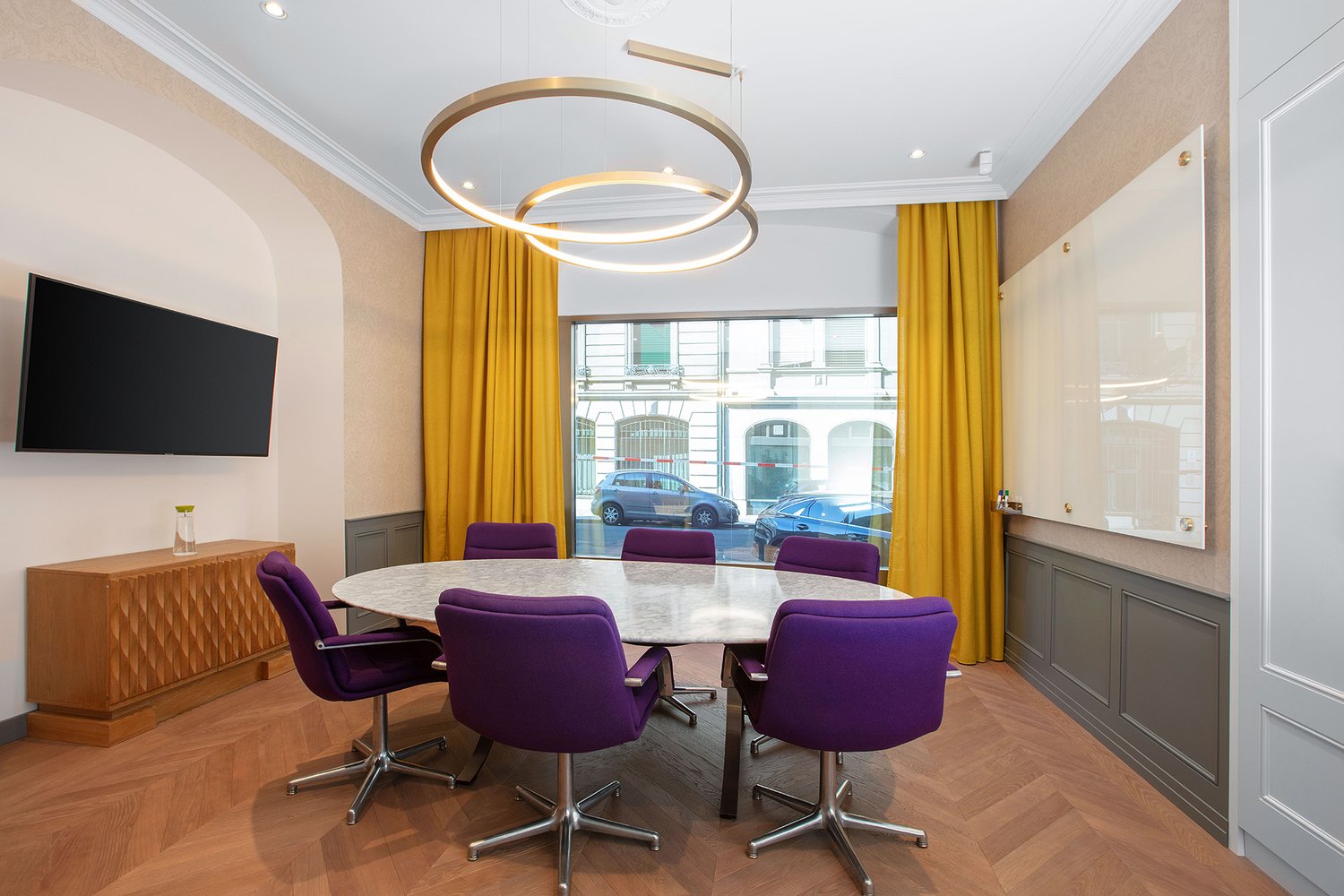 Spacious meeting room with large windows, marble-top dining table, purple upholstered chairs, wooden credenza, and a modern circular lighting fixture. The room has a professional and elegant atmosphere.