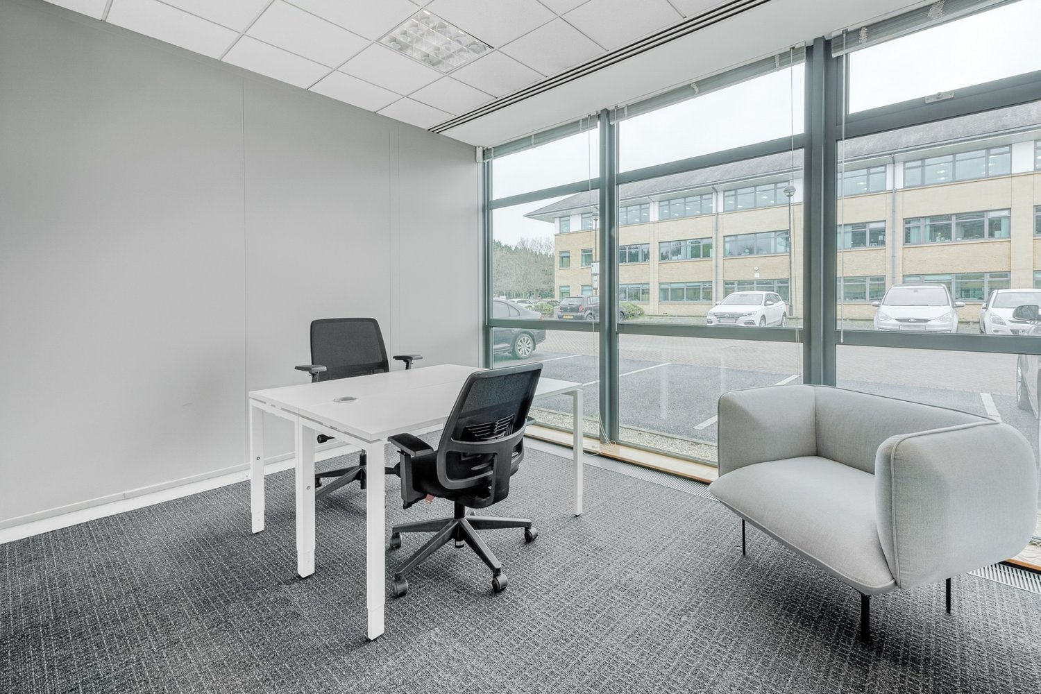 Spacious office with large windows providing a view, white walls, white desk, black office chair, gray sofa, carpeted floor, and ceiling lights