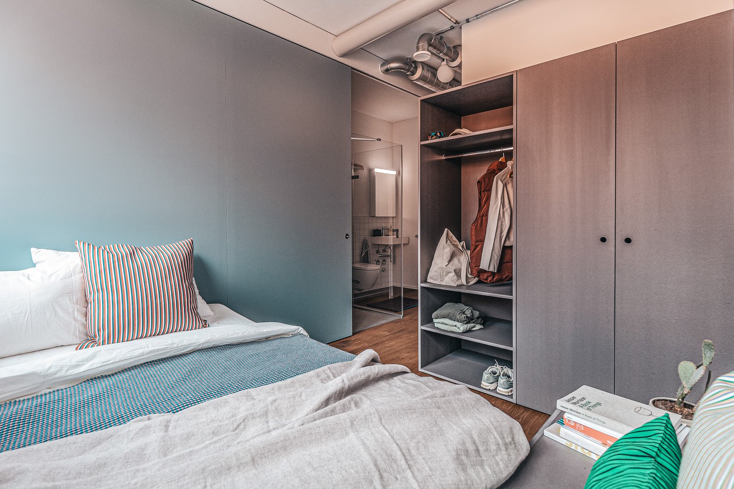 Bedroom with double bed, open door, wooden floor, wardrobe with clothes, bedside table with potted plant, and open bathroom with toilet.