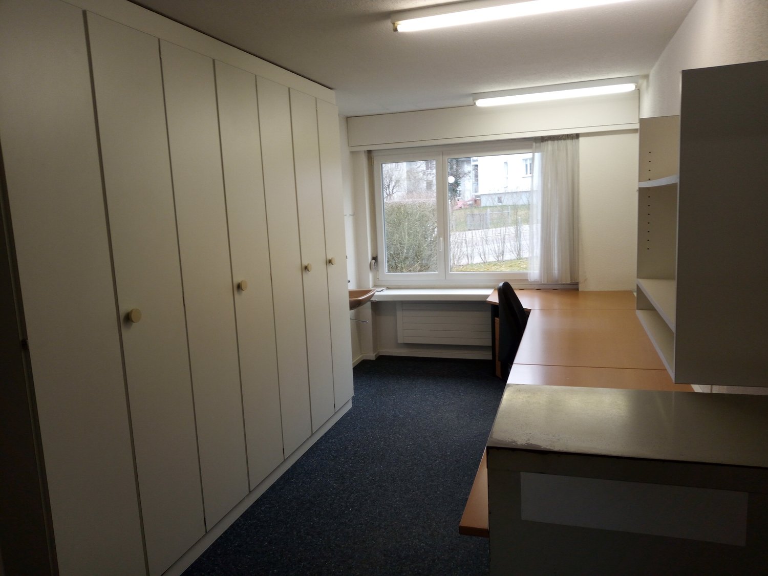 empty room, white cabinets, desk, chair, window, radiator, blue carpet