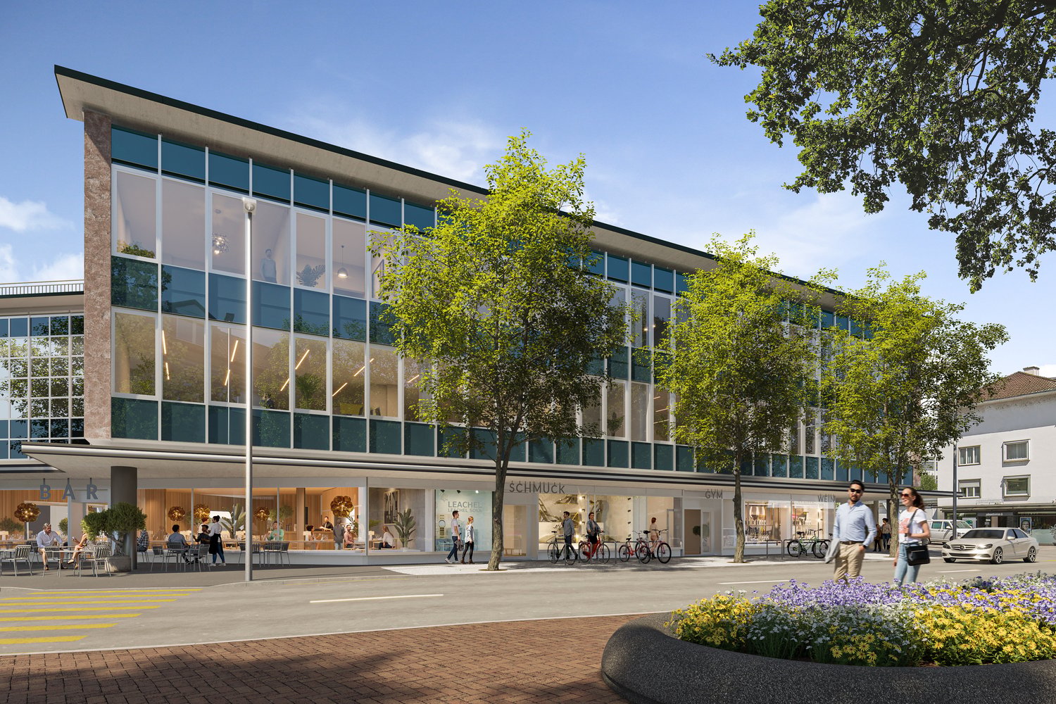 A modern, multi-story commercial building with large glass windows and a prominent entrance. The building has retail stores on the ground floor and appears to be located in a pedestrian-friendly urban area with trees, flowers, and people walking around.