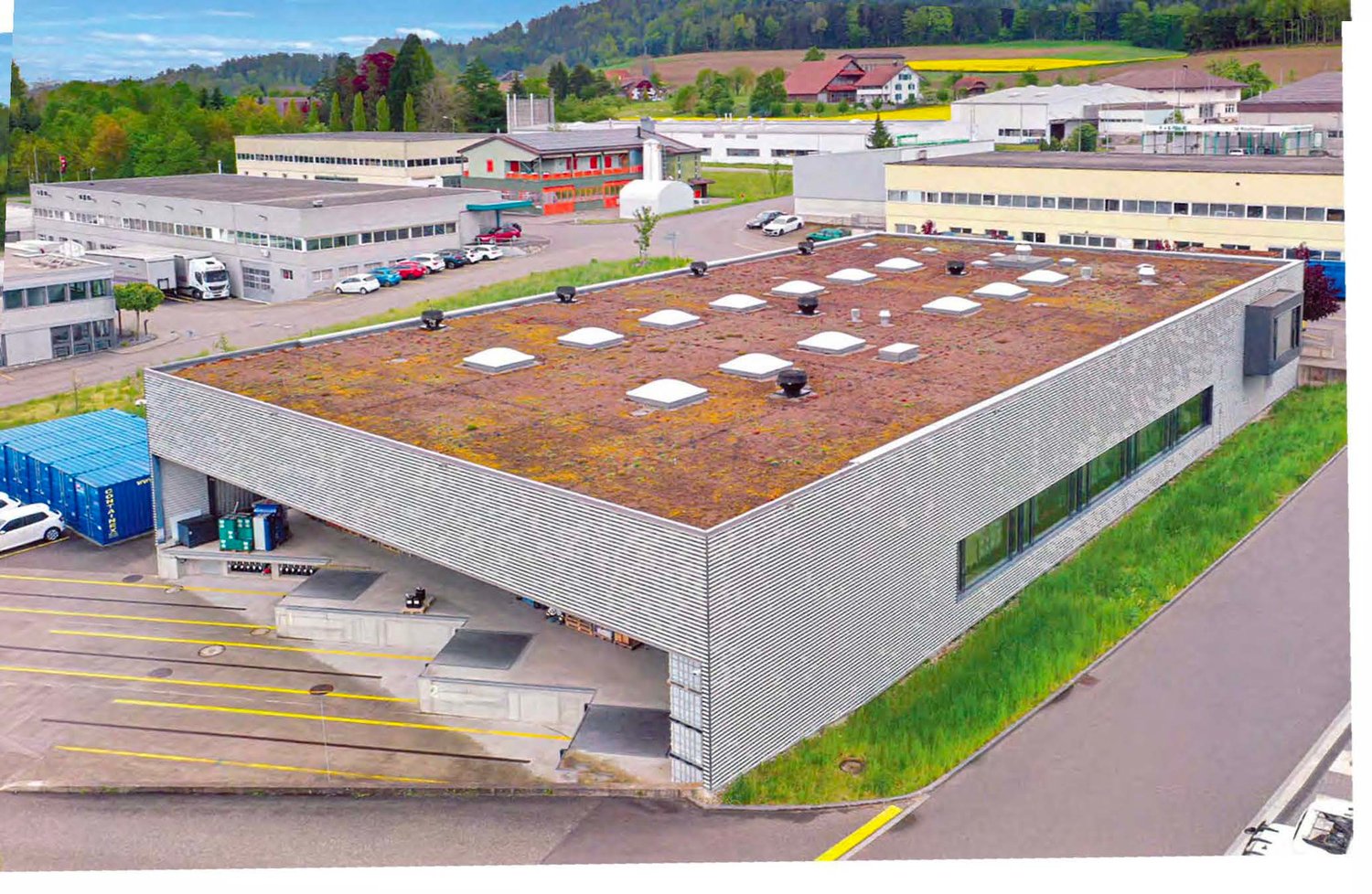 Large commercial building with a green roof, surrounded by parking spaces and other industrial buildings. The building has a modern, industrial design with a flat roof and large windows.