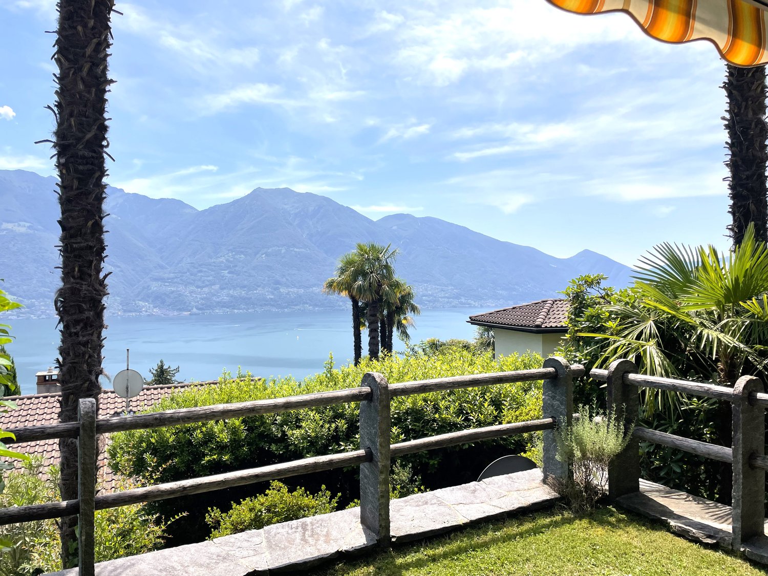 The image shows a scenic outdoor landscape with a view of a lake surrounded by mountains. There is a wooden railing or fence in the foreground, with lush greenery and palm trees in the middle ground. The background features a clear blue sky with some clou