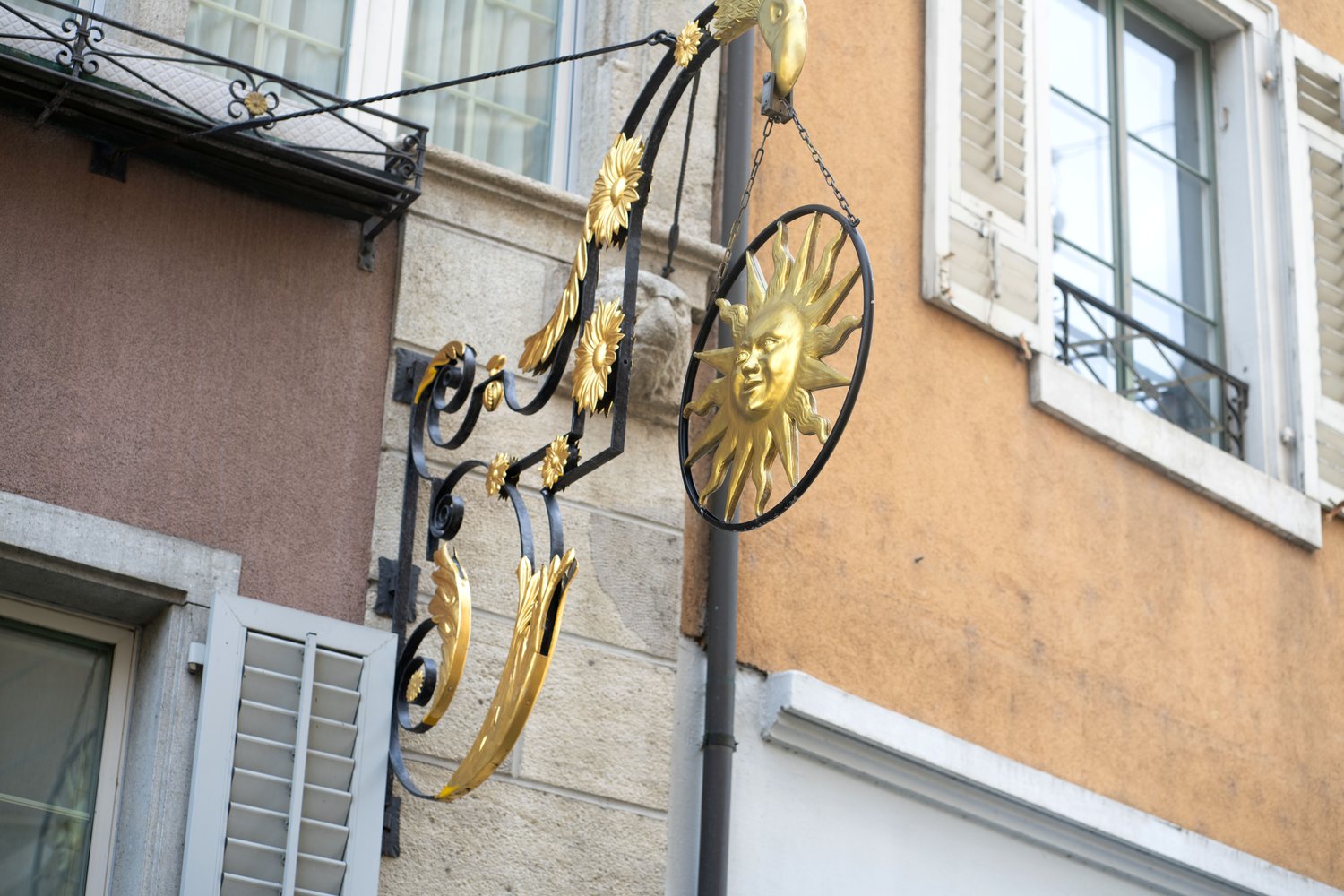 The image shows the exterior of a building with ornate, decorative metalwork featuring golden sun motifs. The metalwork is attached to the facade of the building, which has large windows and a textured, beige exterior. There appears to be a balcony or ter