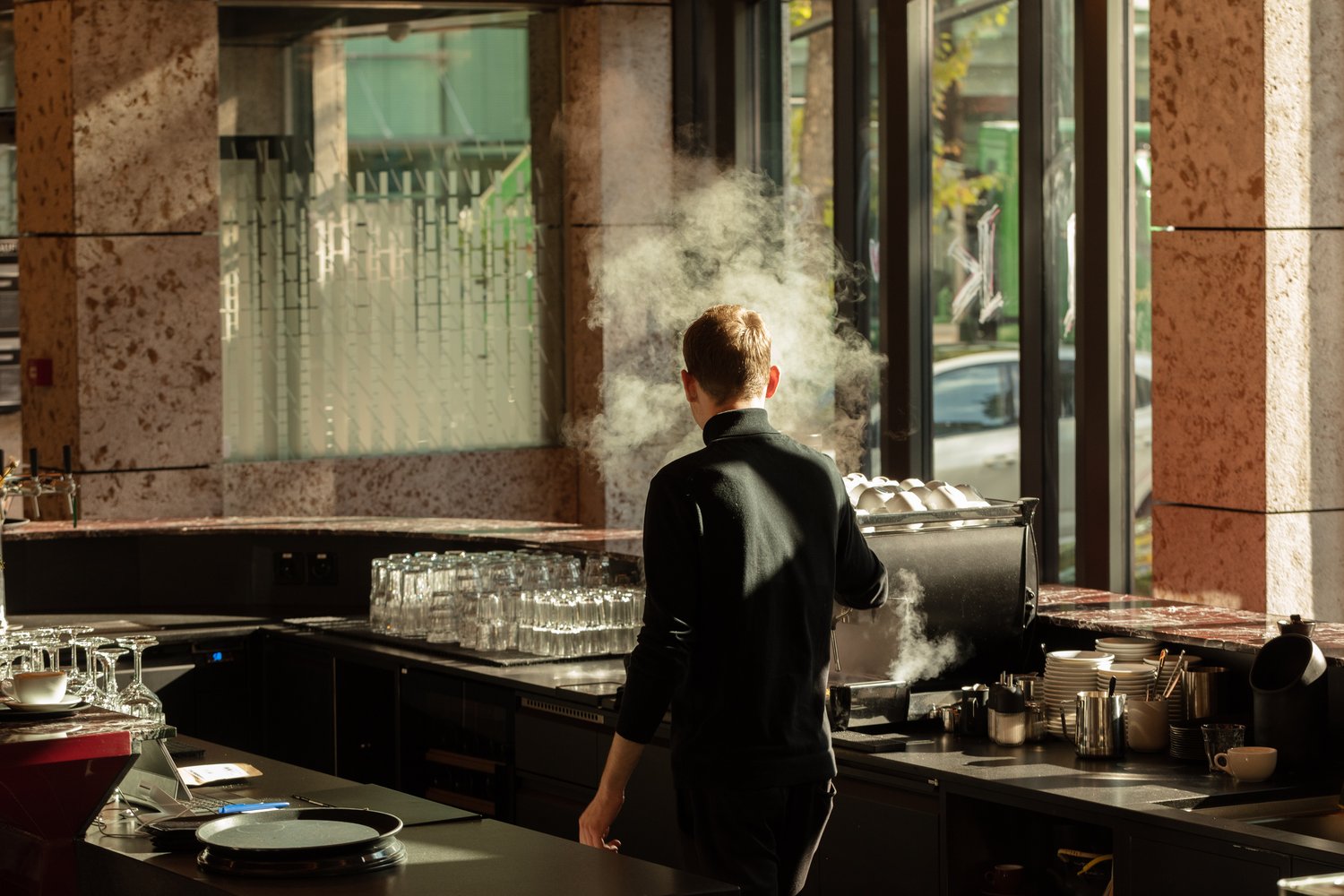 The image shows the interior of a restaurant kitchen. A chef in a black uniform is standing in front of a large commercial kitchen appliance, with steam or smoke visible. The kitchen appears to be well-equipped with various cooking utensils, dishes, and g