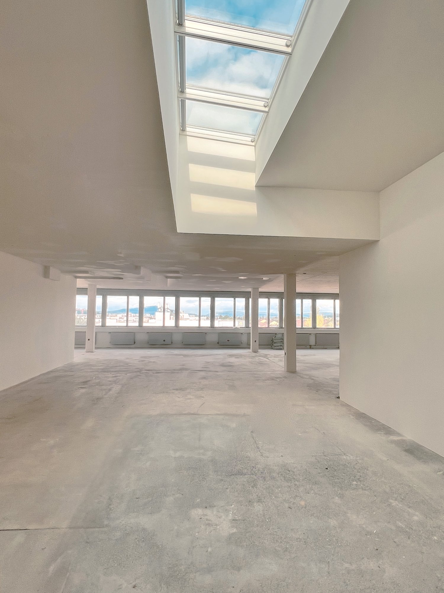 Large open-plan office space with concrete floor, white walls, and large windows providing a view of the surrounding area. The space has a high ceiling with a skylight, allowing natural light to flood the room.