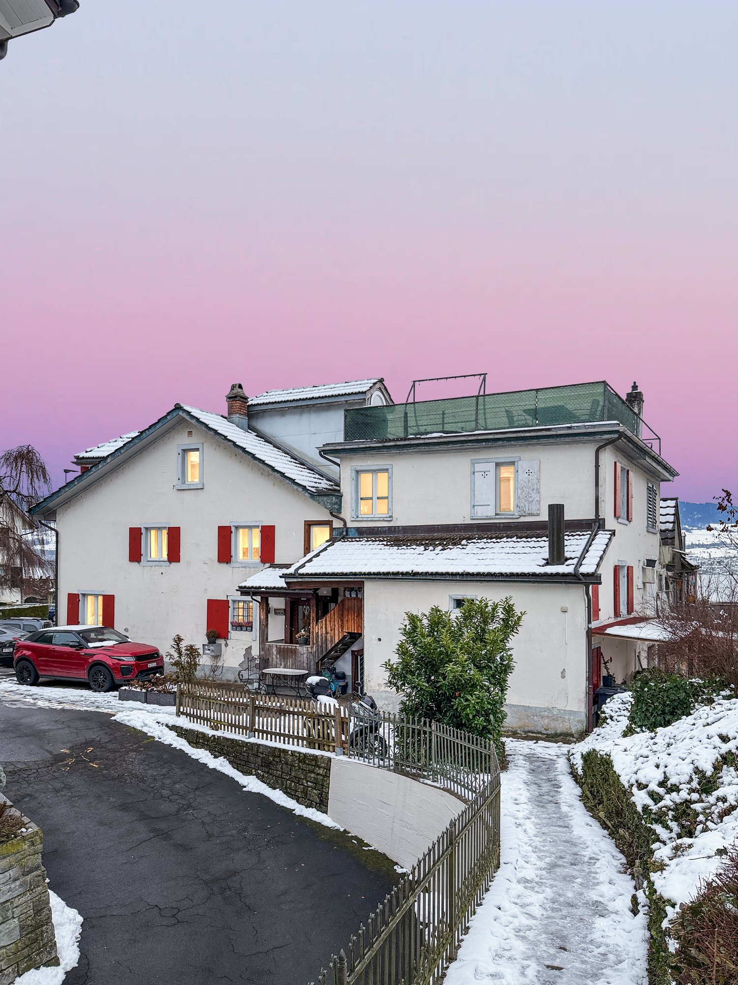 2 story house, white exterior, red shutters, snow on the roof, red car parked in front, pathway leading to the entrance, small garden with plants, fence, and a small wooden structure