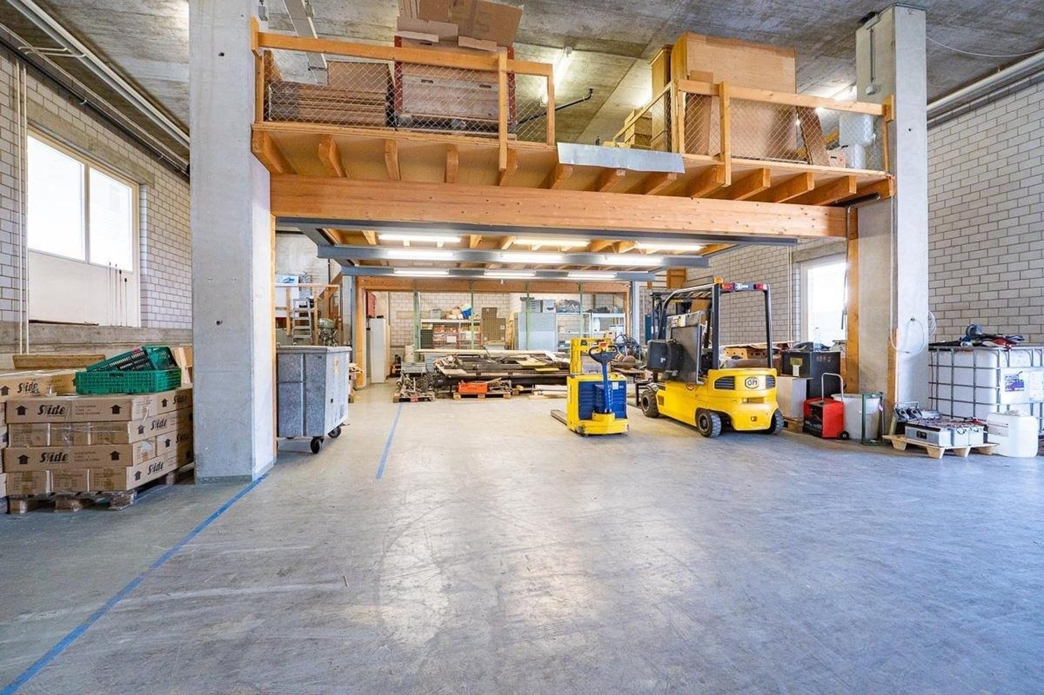 Empty warehouse with a large open area, wooden mezzanine with storage boxes, a few stacks of wooden pallets with boxes, two forklifts, machinery, metal barrels, and containers