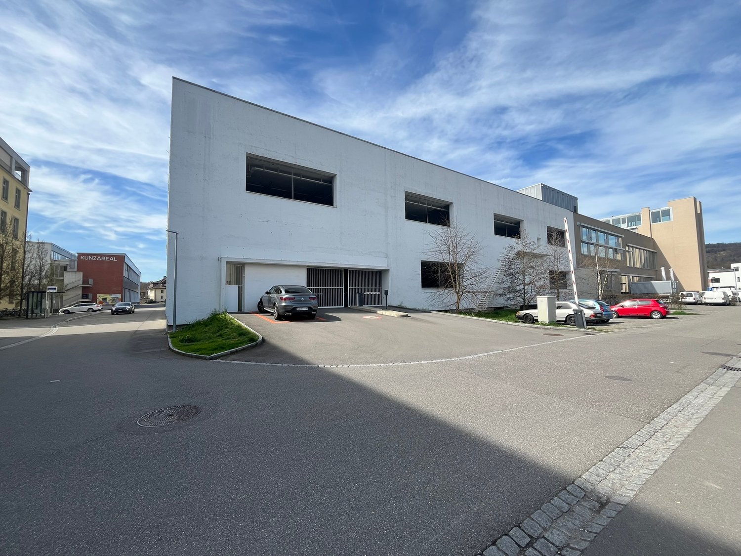 A large, modern commercial building with a flat roof and multiple windows. There is a parking area in front of the building with several cars parked. The building appears to be used for commercial or industrial purposes.