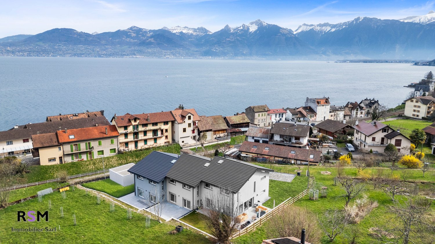 The image shows a residential area with a mix of multi-story buildings and single-family homes. The buildings have traditional alpine-style architecture with red tile roofs. The homes are surrounded by lush green lawns and trees. In the background, there