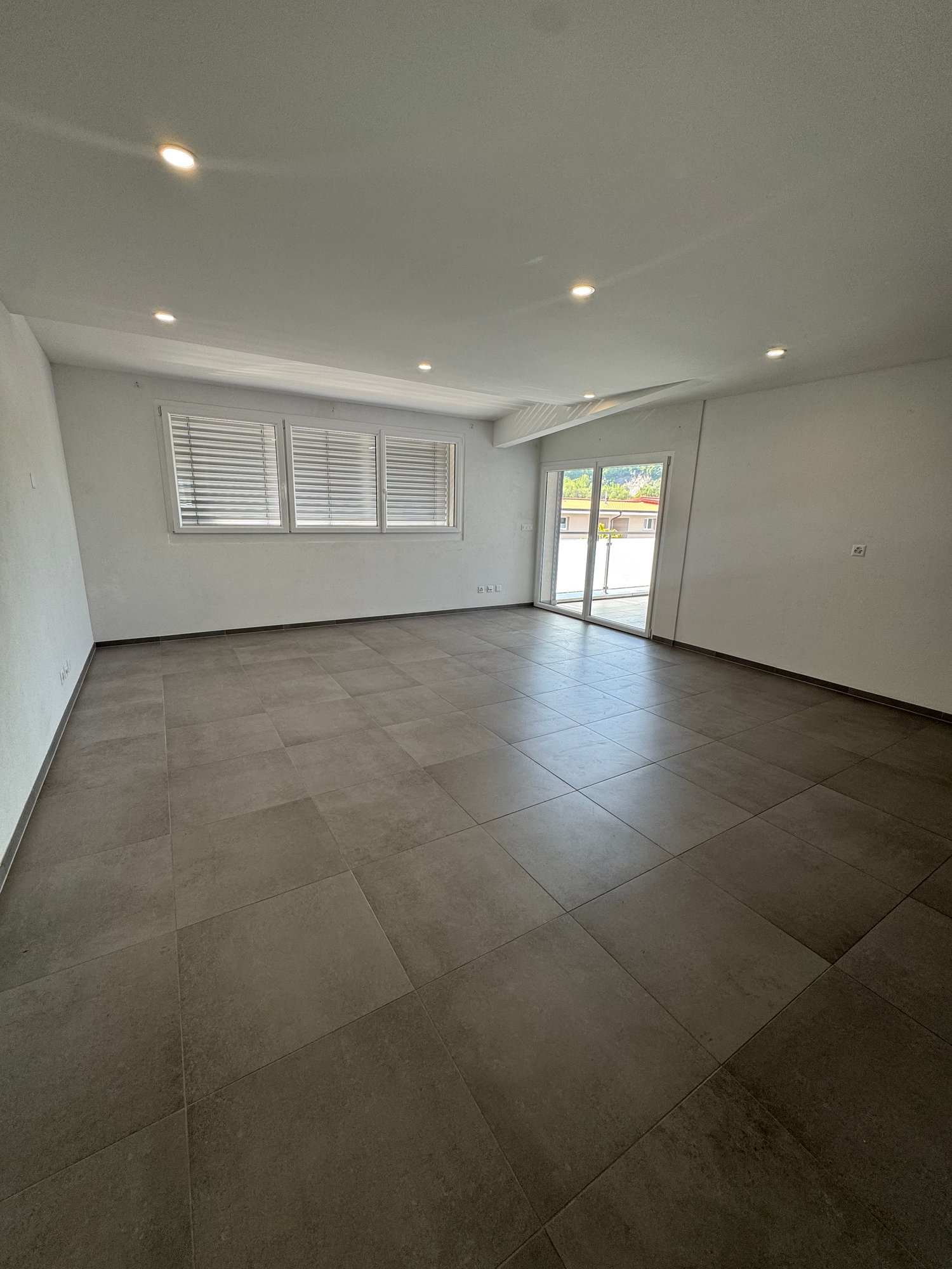 Spacious living room with large windows, gray tile flooring, and recessed lighting. The room has access to a balcony or terrace through sliding glass doors.
