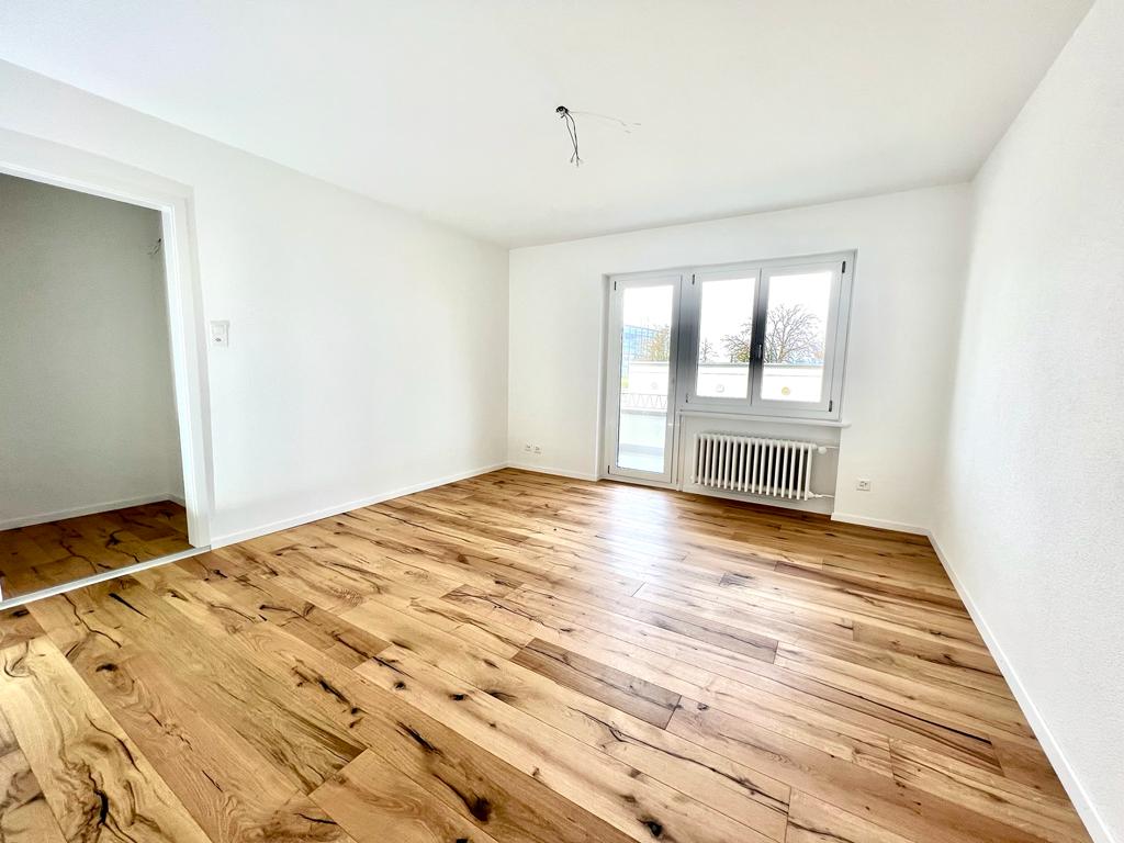 An empty room with hardwood floors and white walls. The room has a glass door leading to a balcony. There is a radiator below the balcony window.