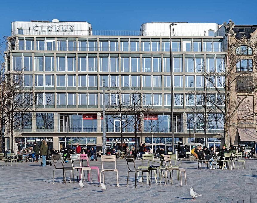 Modern office building with glass facade, multiple floors, outdoor seating area, people walking around, trees, street lights, clear sky