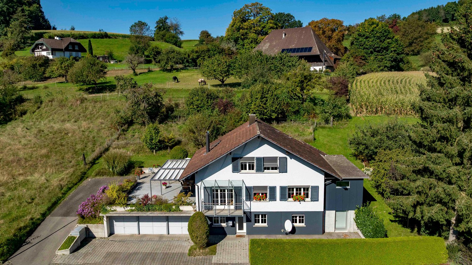 Two-story house with brown roof, white walls, large windows, garage, garden, and a covered terrace.