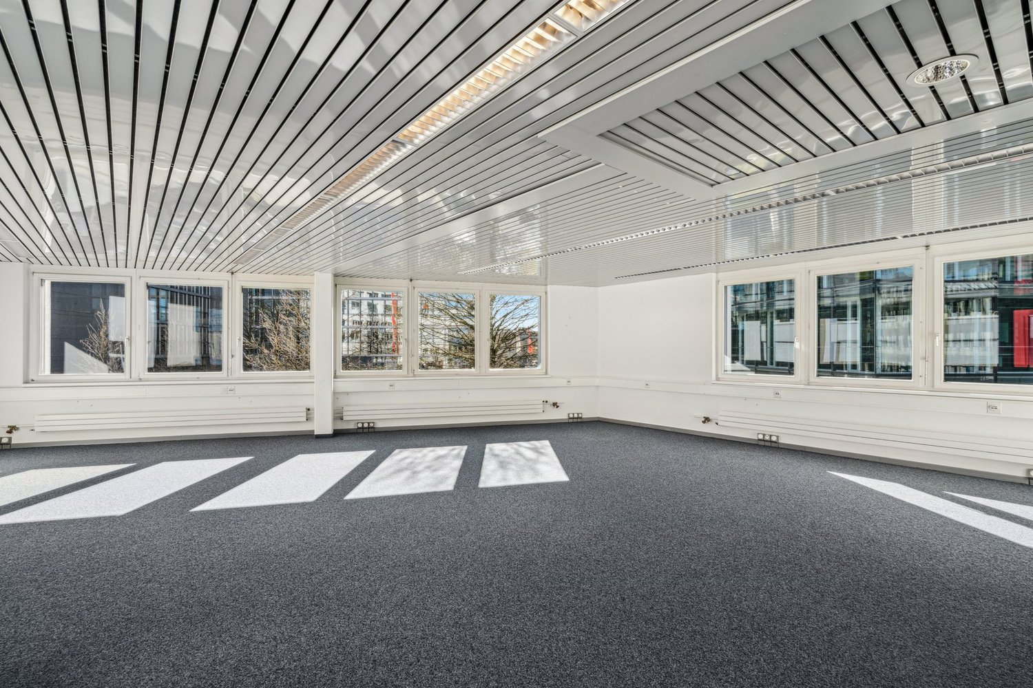 A large empty room with carpeted floor, multiple large windows, metal ceiling panels, recessed lighting, and no furnishings