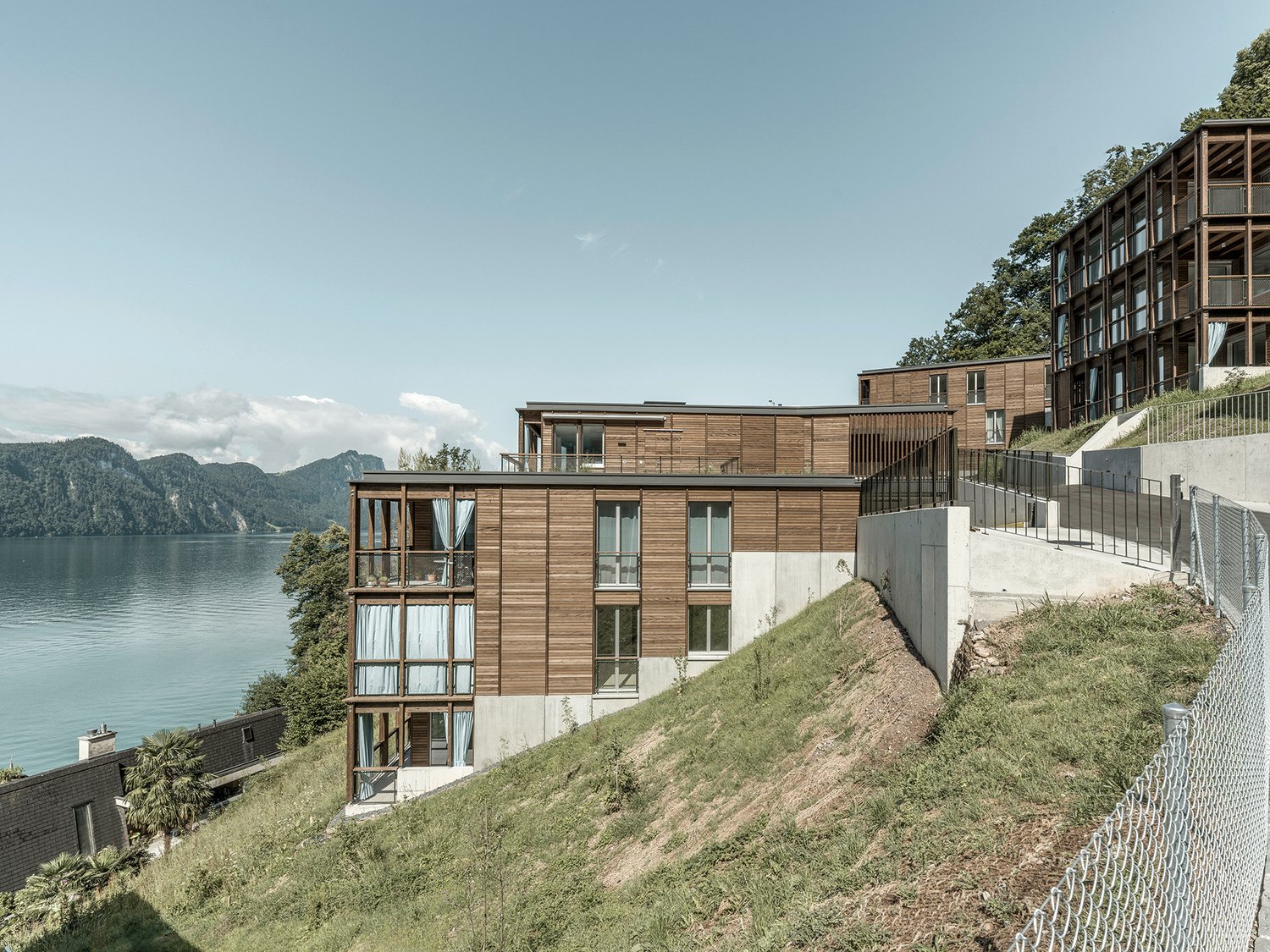 Multi-story apartment building with wooden exterior, large windows, and balconies overlooking a lake and mountains in the background