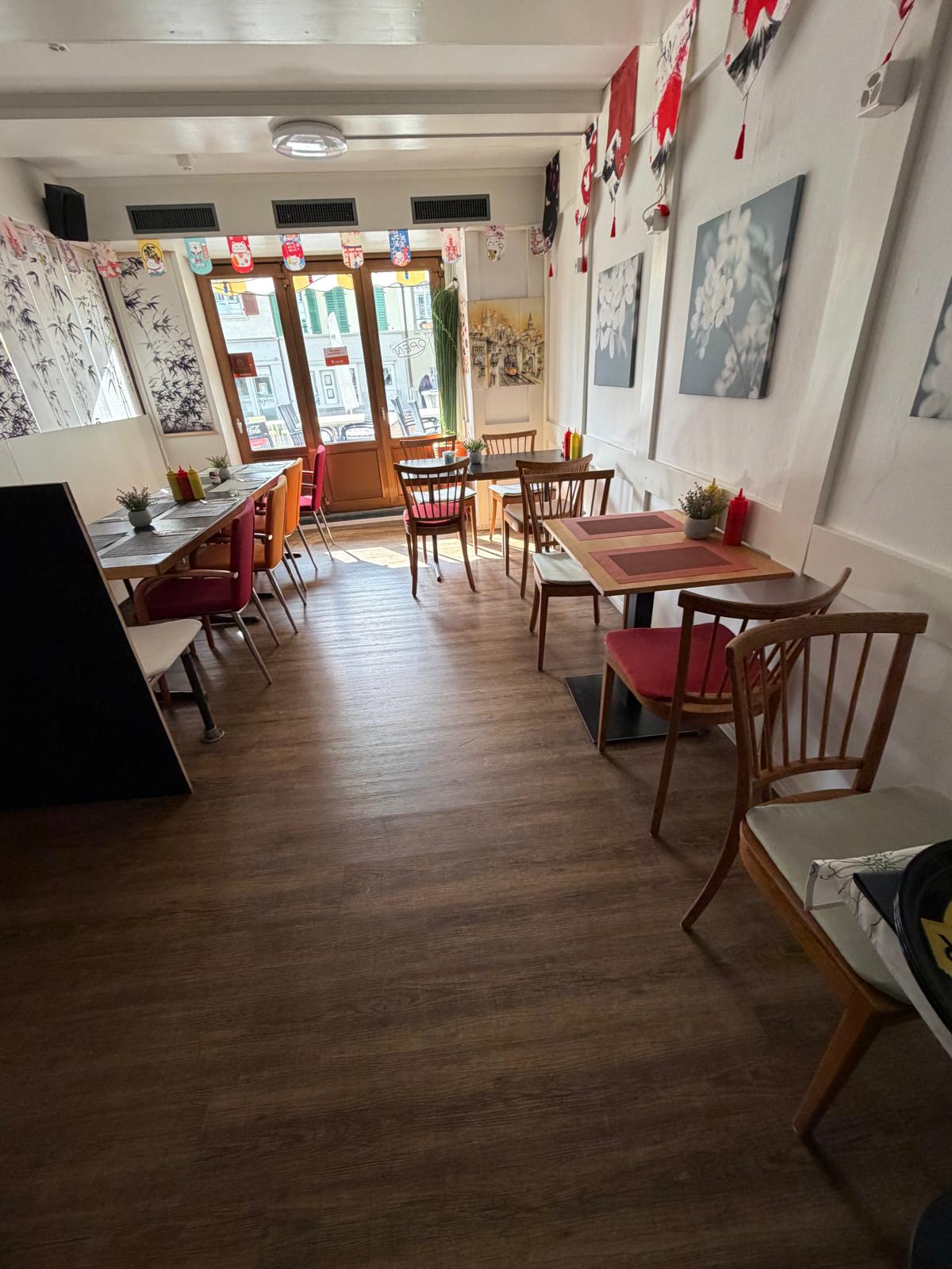 empty restaurant with wooden floor, wooden tables and chairs, white walls, decorative items, and a glass door leading to the outside